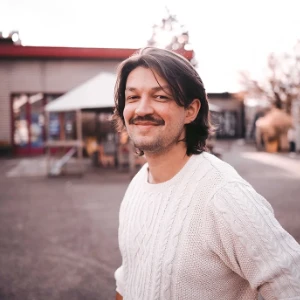 Smiling man with mustache and shoulder-length hair wearing a white knit sweater outdoors.