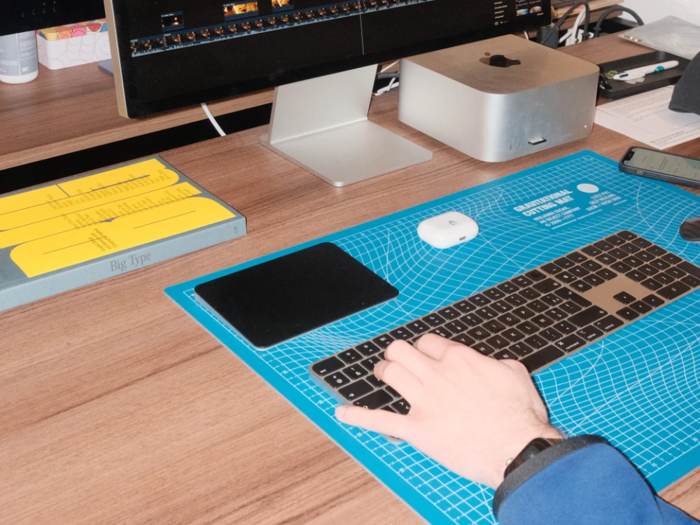 Saisie à la main sur un clavier sans fil posé sur un tapis de découpe bleu, à côté d’un Mac Studio et d’un moniteur sur un bureau.