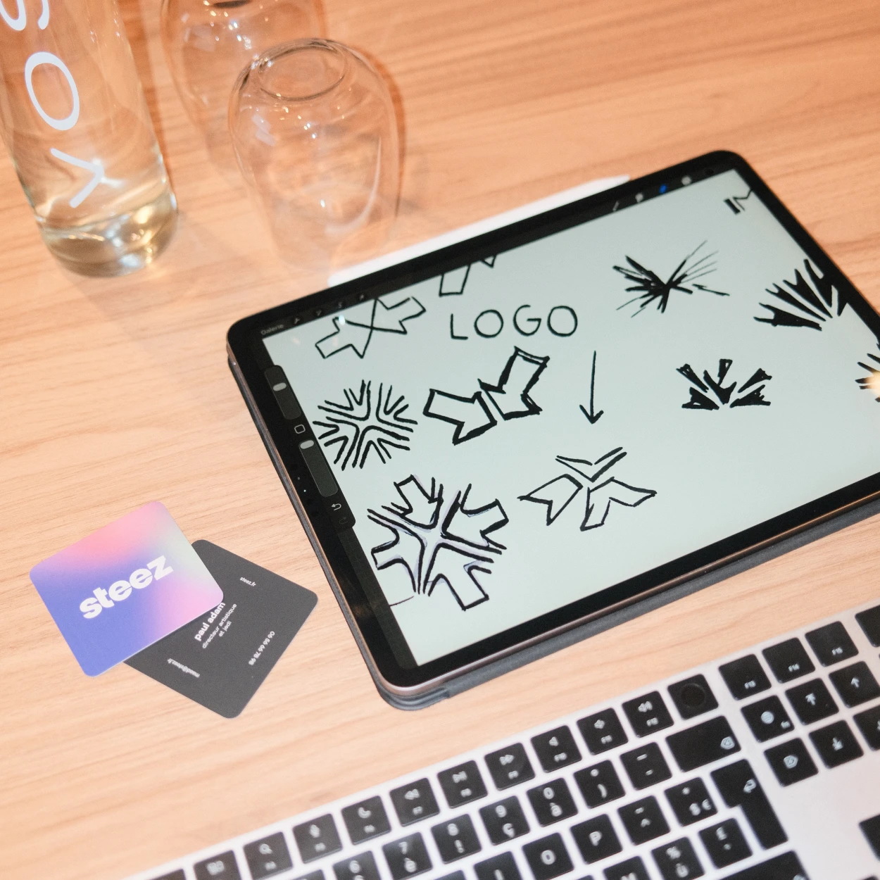 Tablette sur le bureau affichant des esquisses de logo noir avec une flèche près du clavier, deux cartes et une bouteille en verre.