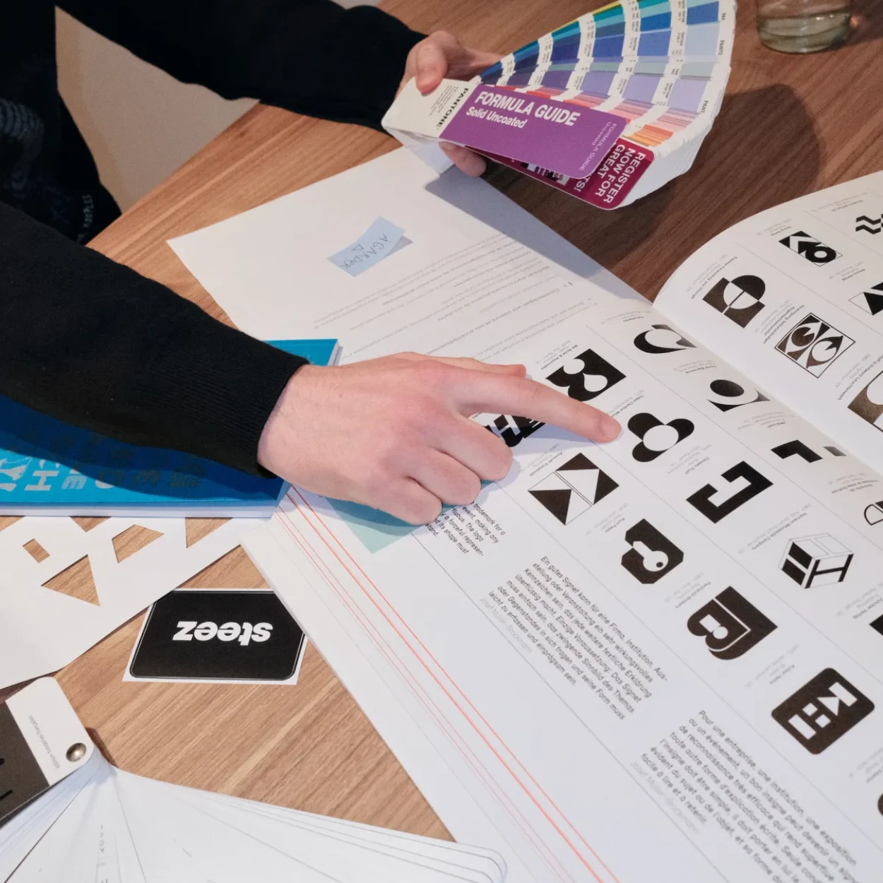 Mains examinant un catalogue de logos en noir et blanc avec un nuancier de couleurs sur une table en bois.
