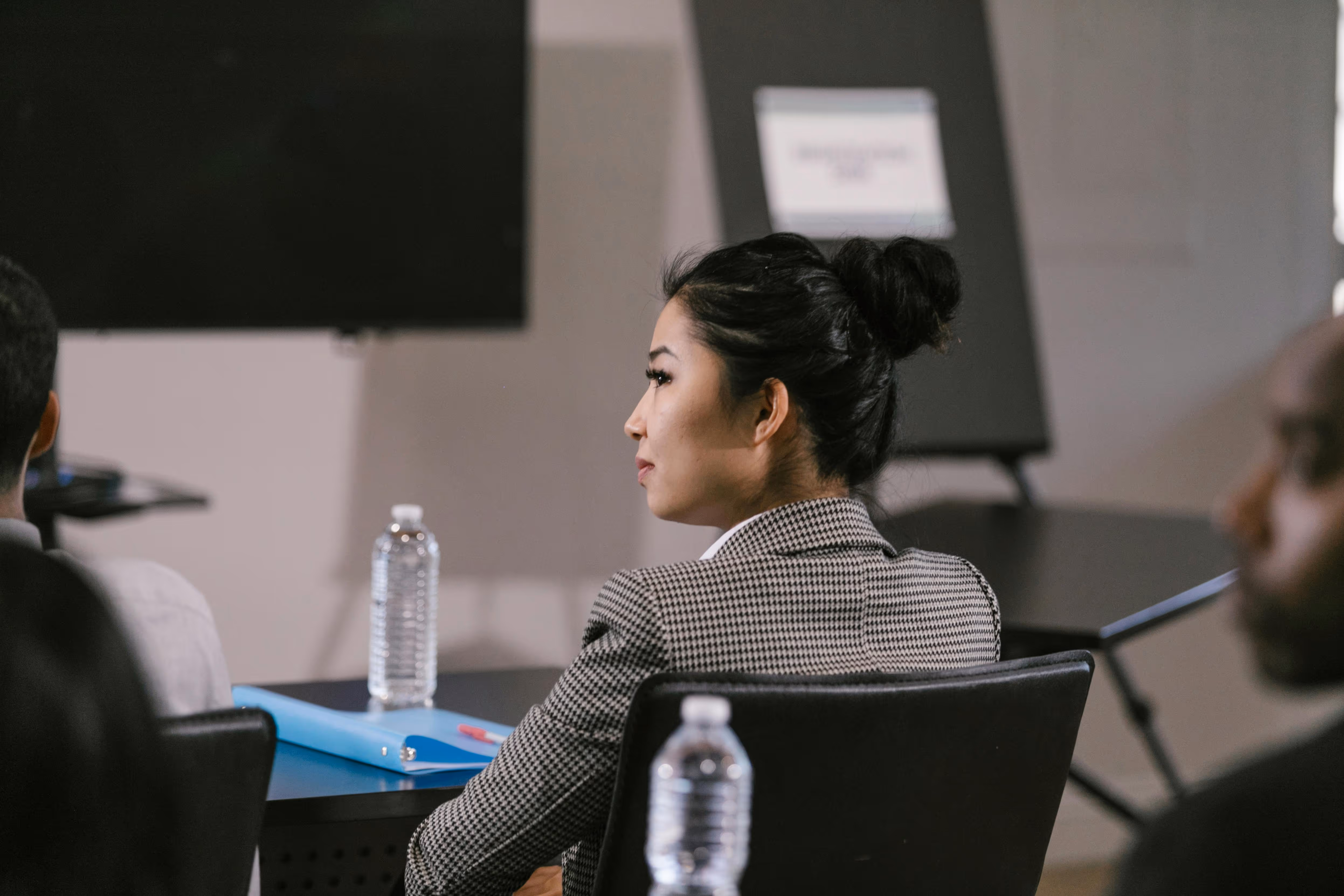 Woman with black hair tied in a bun, wearing a houndstooth blazer, sitting attentively in a conference room.