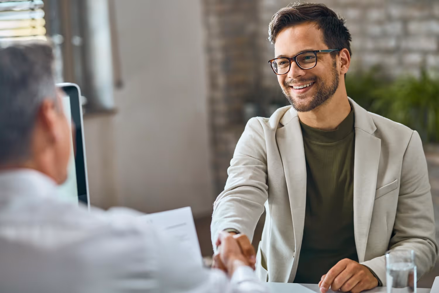 Smiling man with glasses shaking hands with another person at a desk with a glass of water.