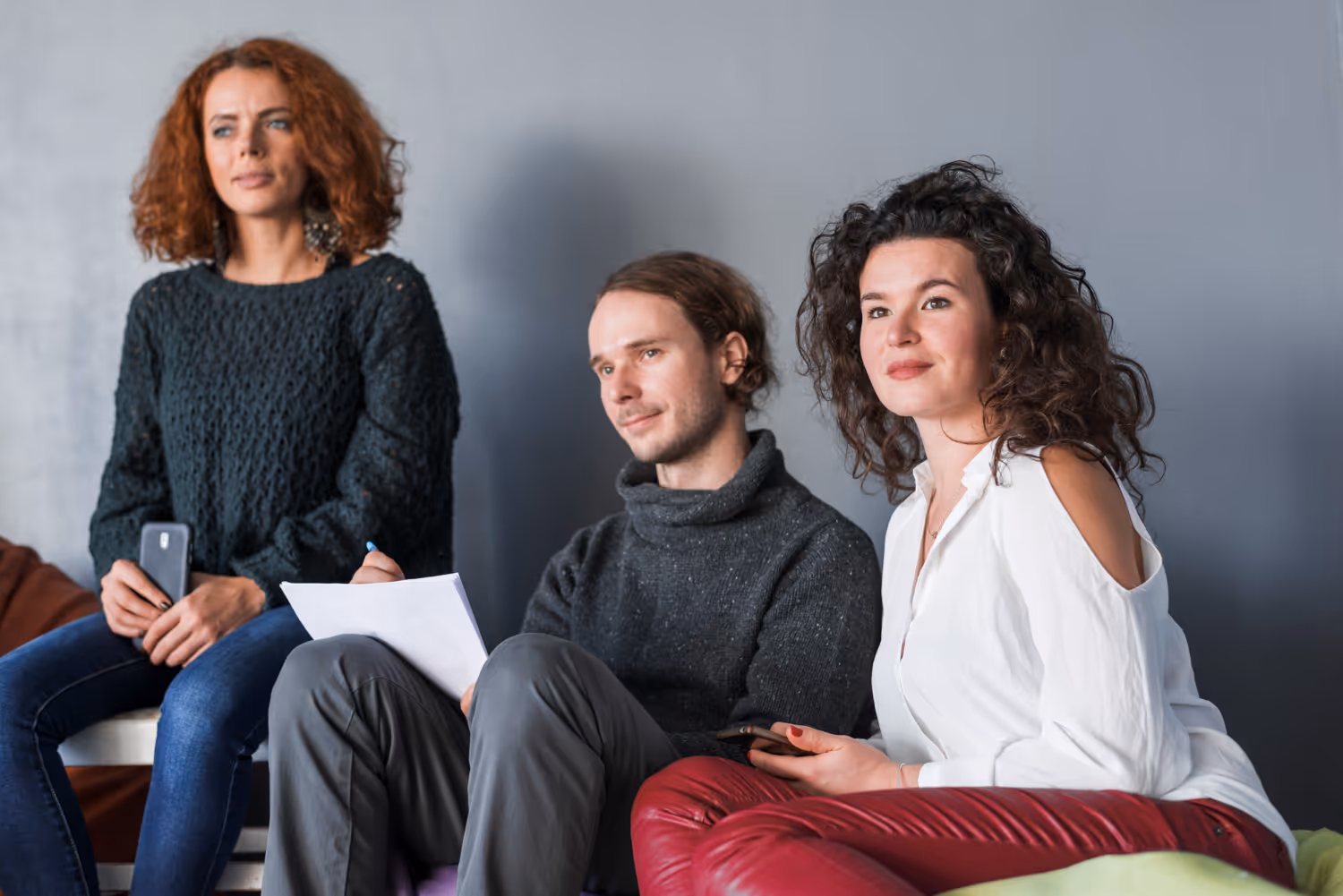 Three young adults sitting against a gray wall; one woman with curly red hair in a dark sweater holding a phone, a man in a gray sweater with a notepad, and a woman with curly dark hair in a white blouse and red pants.