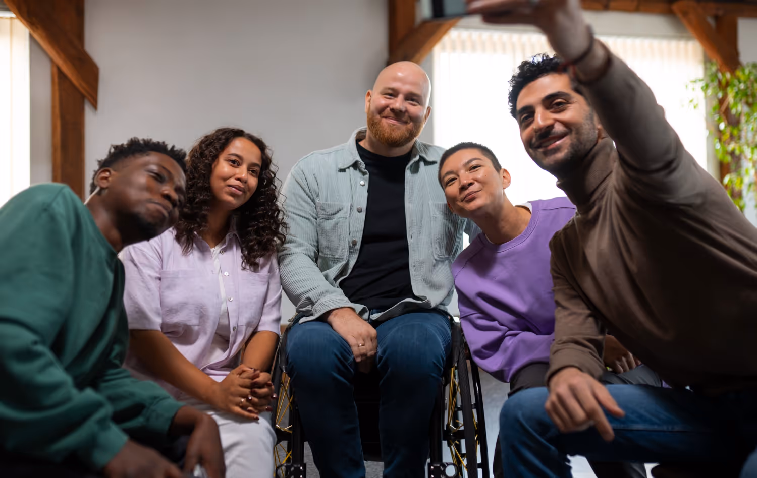 Diverse group of five smiling friends, including a man in a wheelchair, taking a selfie indoors.