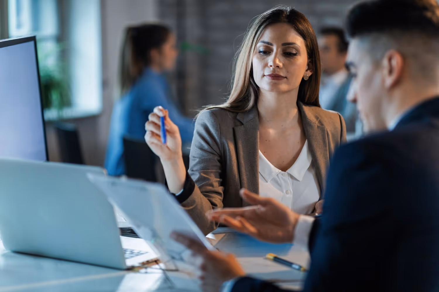 Businesswoman holding a pen and discussing documents with a colleague in an office setting.