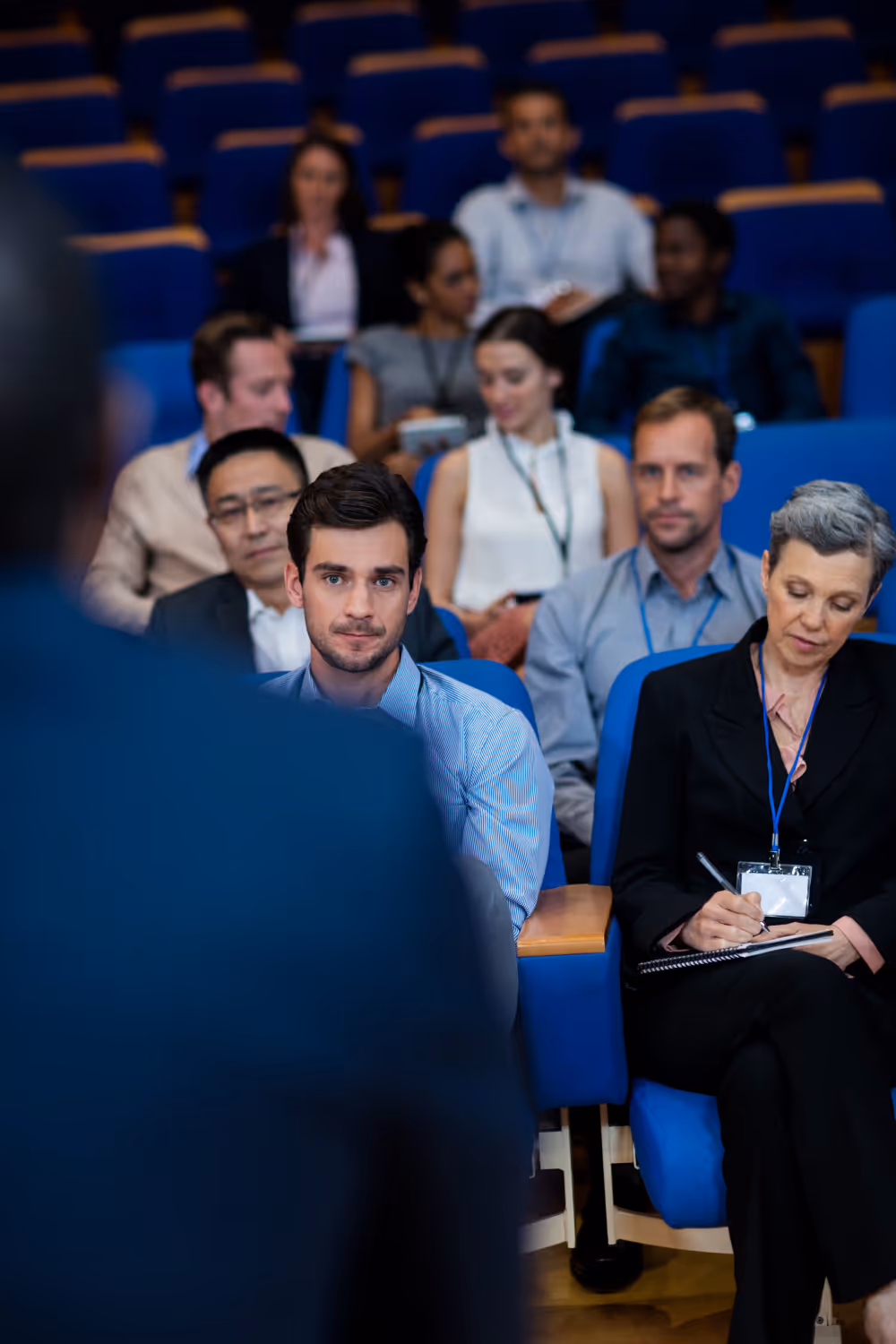 Audience members seated in a lecture hall listening to a speaker, with one woman taking notes.
