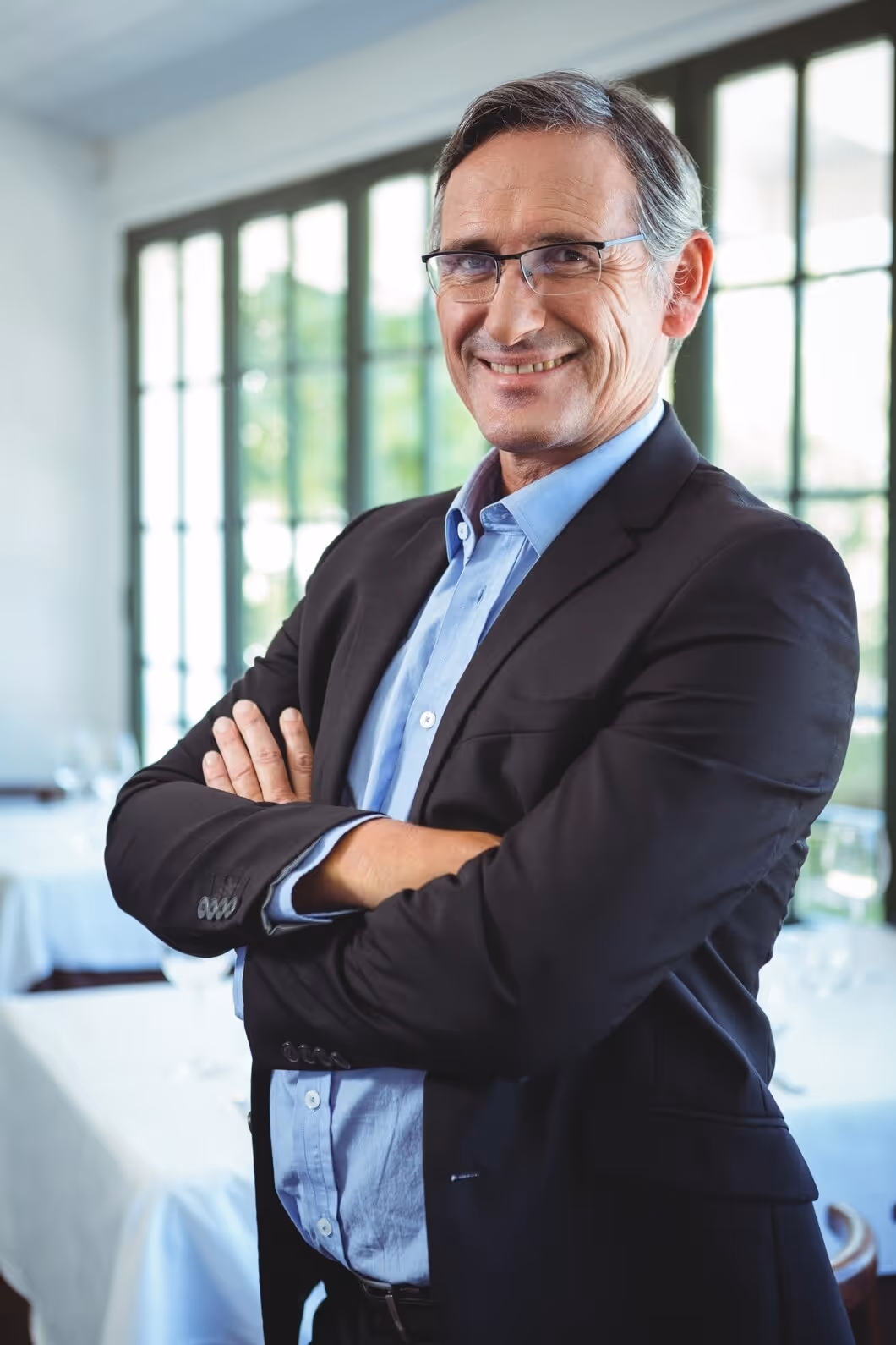Smiling middle-aged businessman wearing glasses, a blue shirt, and black blazer standing with arms crossed in a bright room with large windows.