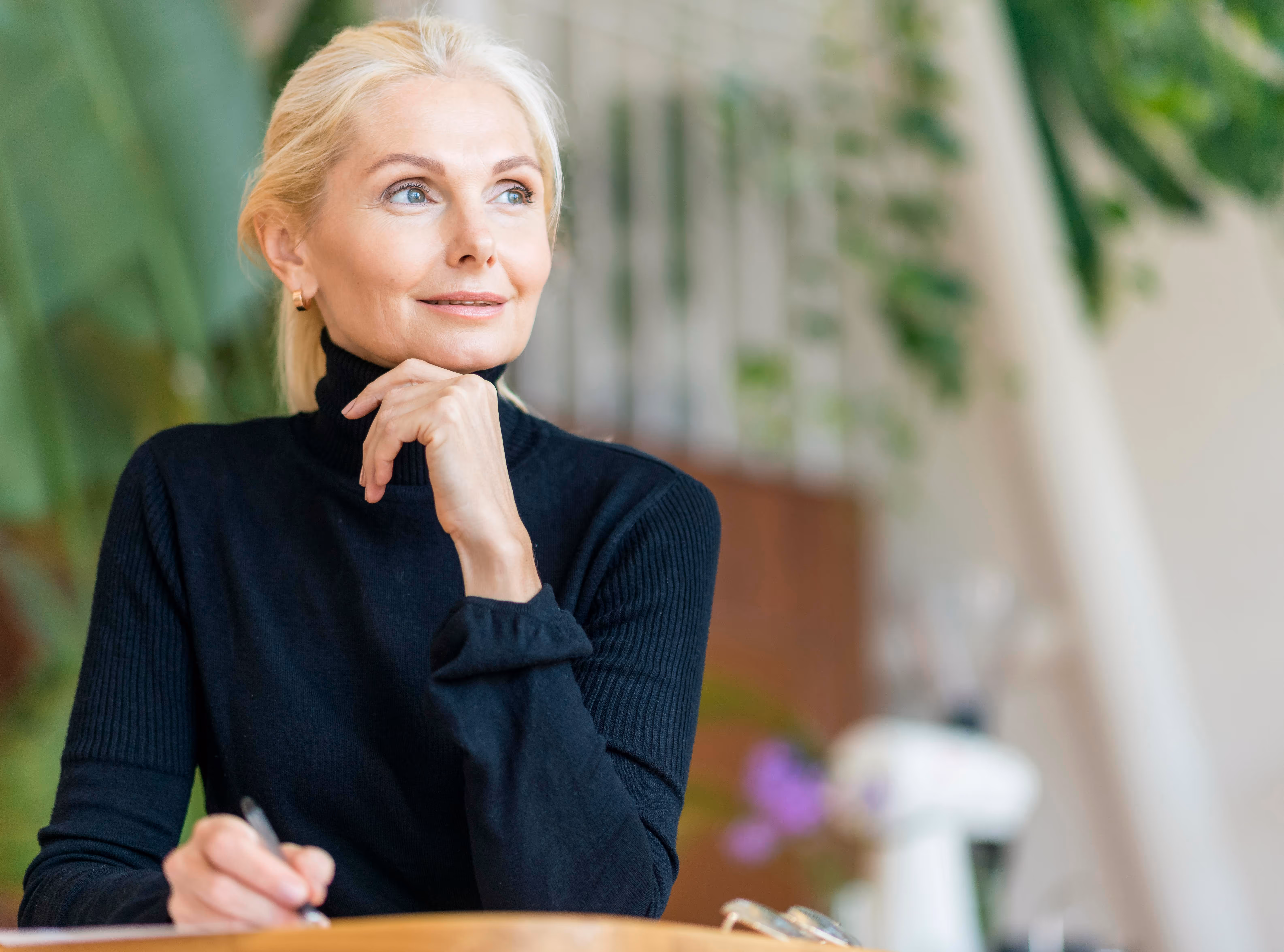Older woman with blonde hair wearing a black turtleneck, holding a pen and looking thoughtfully to the side.