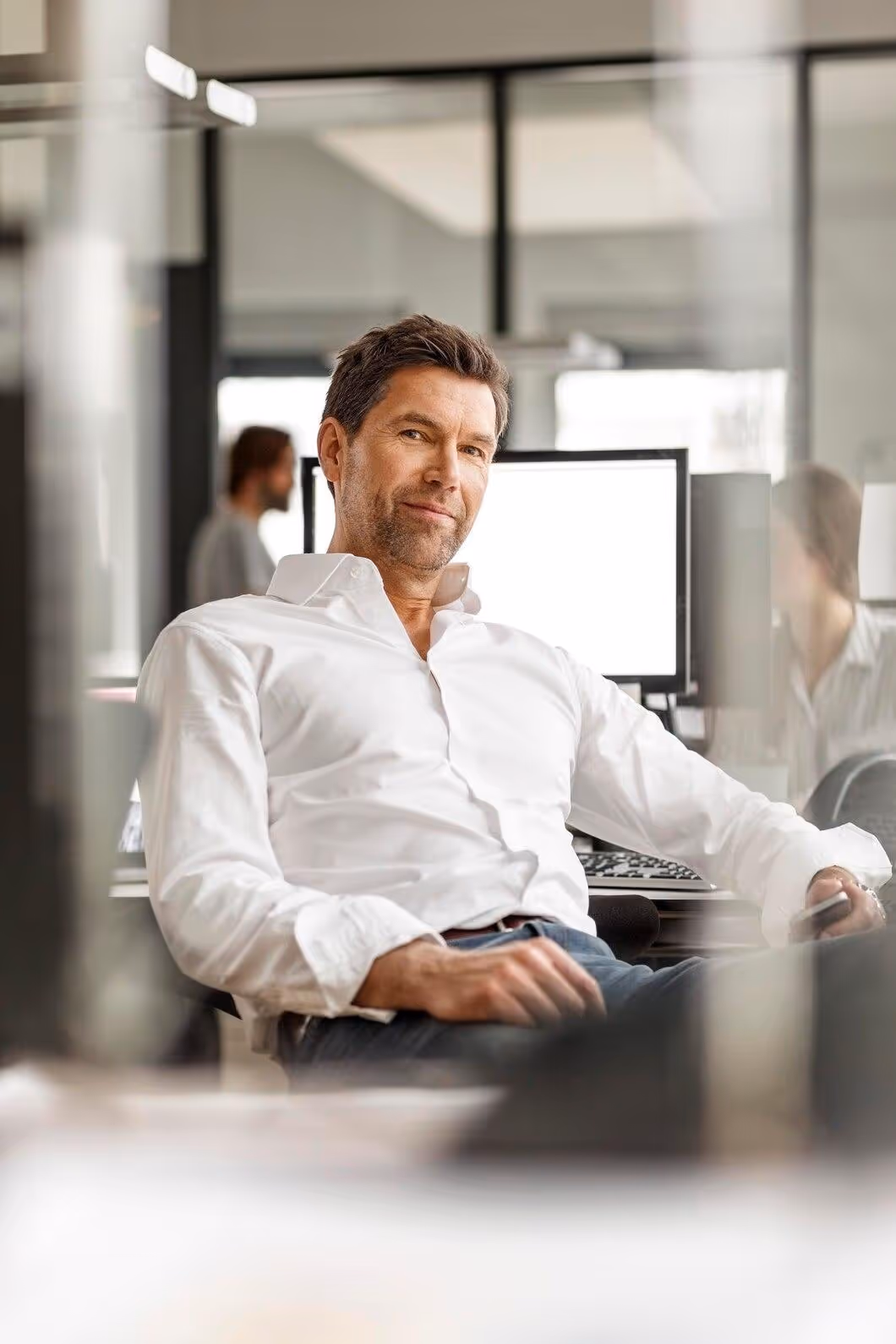 Confident businessman in a white shirt sitting relaxed at his office desk with a computer monitor behind him.