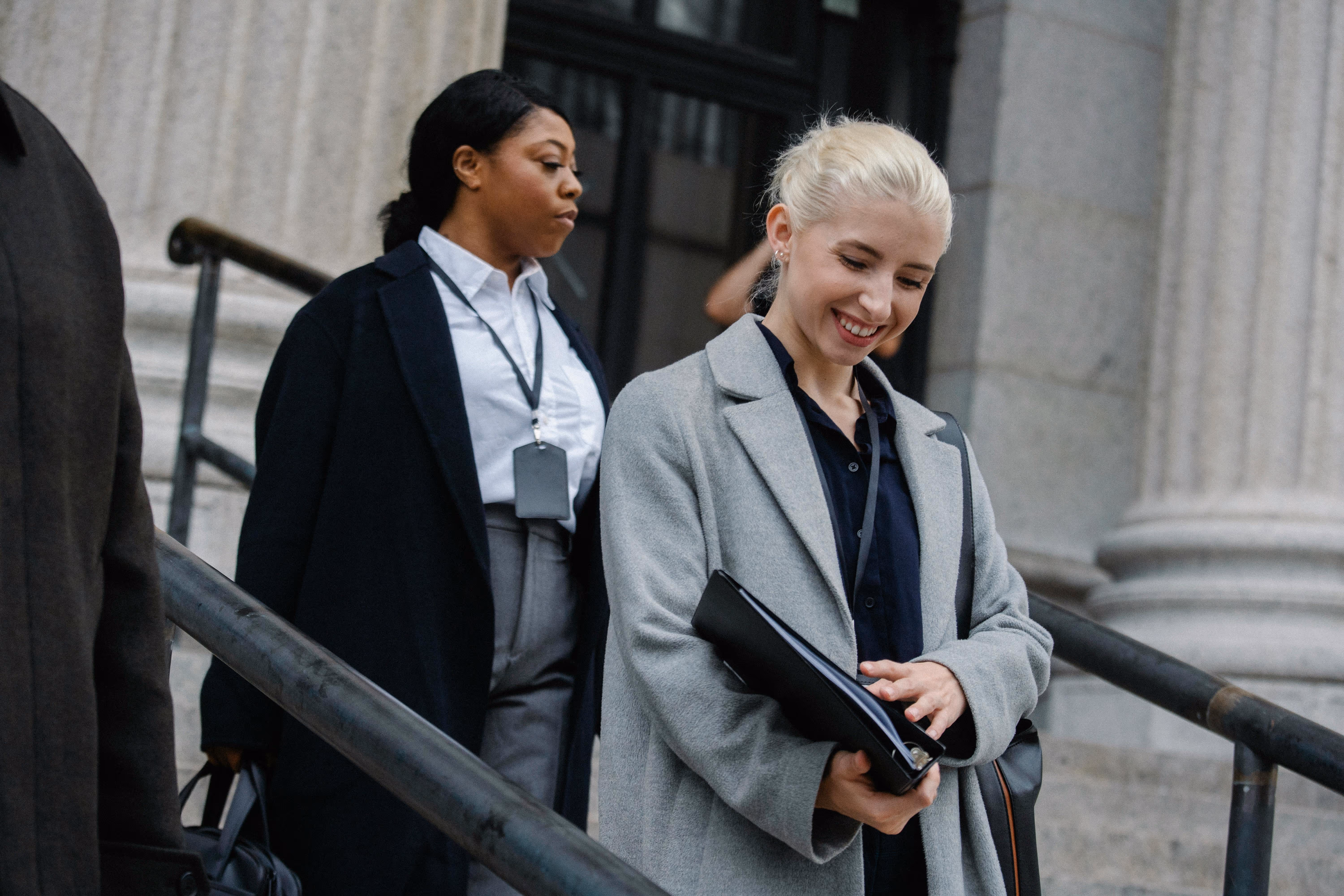 Two businesswomen descending stone steps outside a classical building, one smiling and holding a folder.