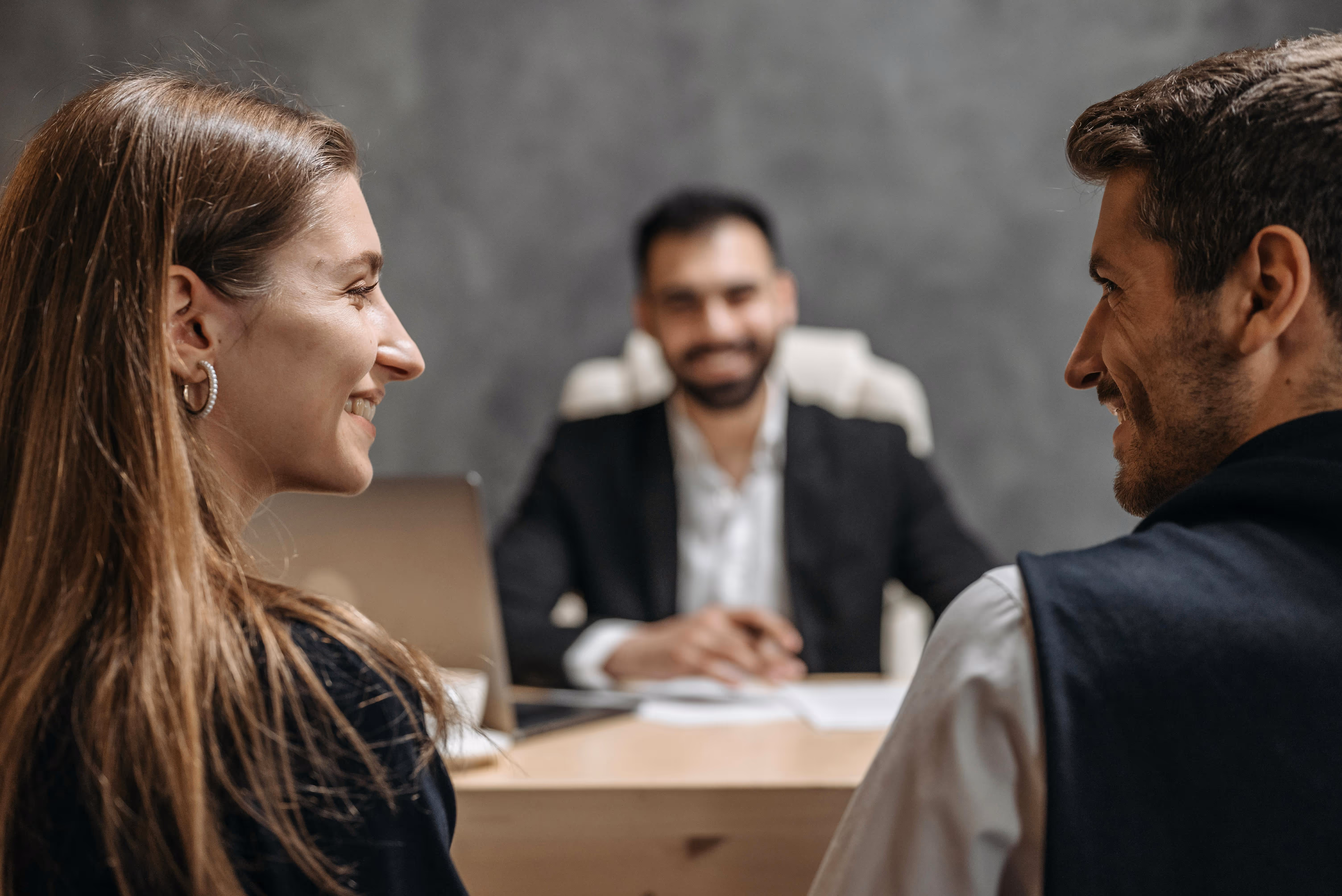 Smiling woman and man facing each other with a man in a suit sitting at a desk in the background.