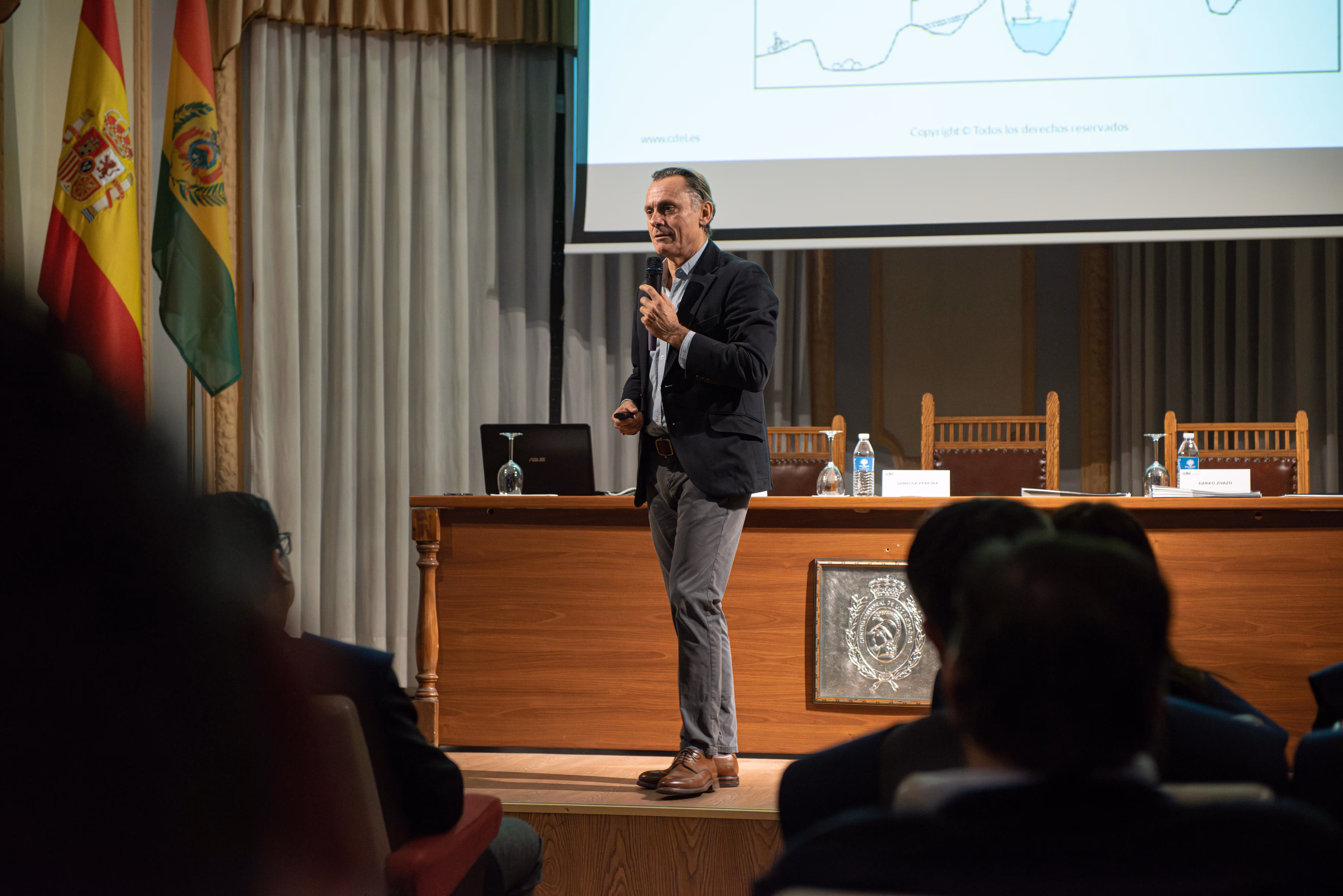 Man in business attire giving a presentation on stage with Spanish and Bolivian flags in the background and an audience watching.