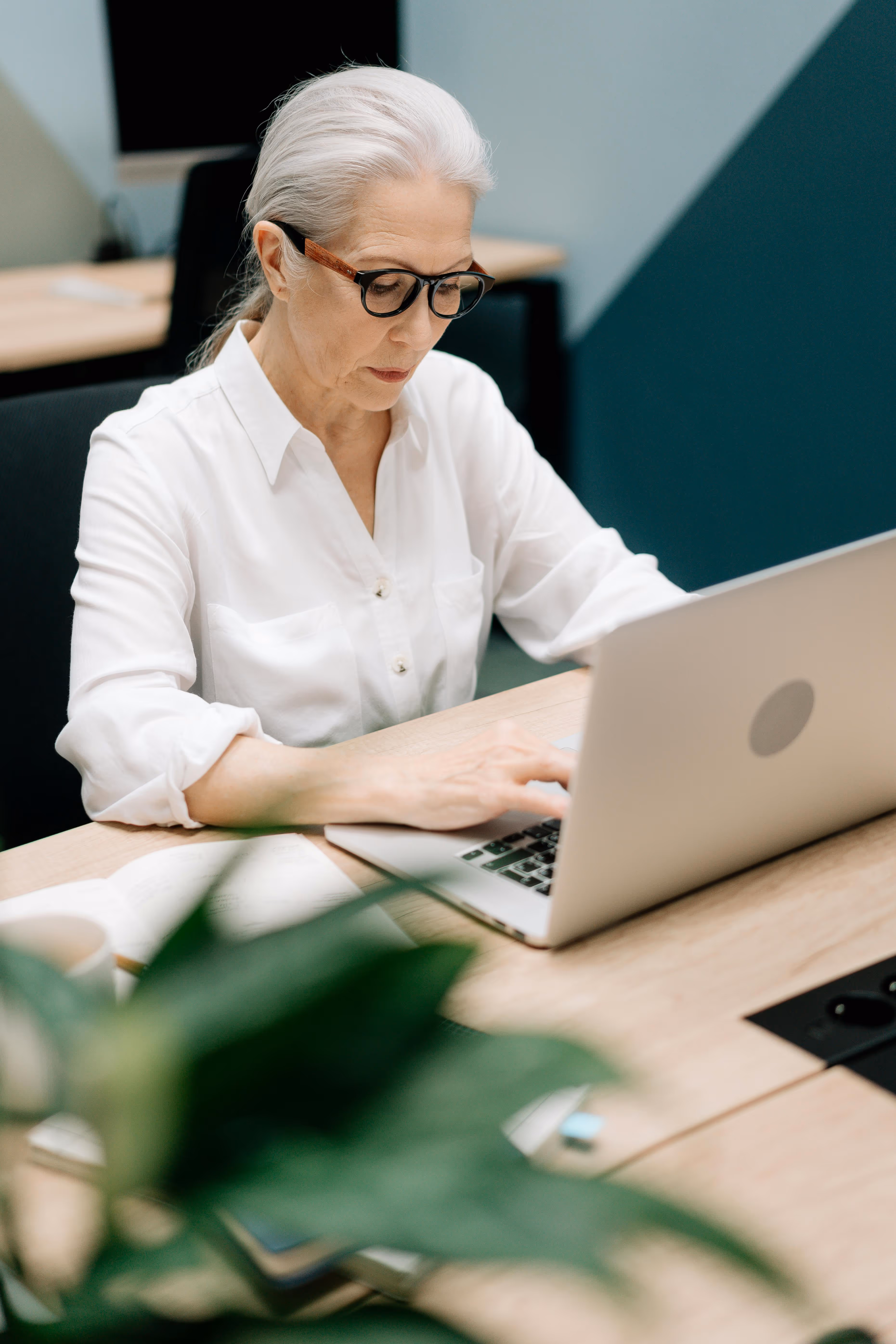 Older woman with gray hair and glasses working on a laptop at a wooden desk in a modern office.