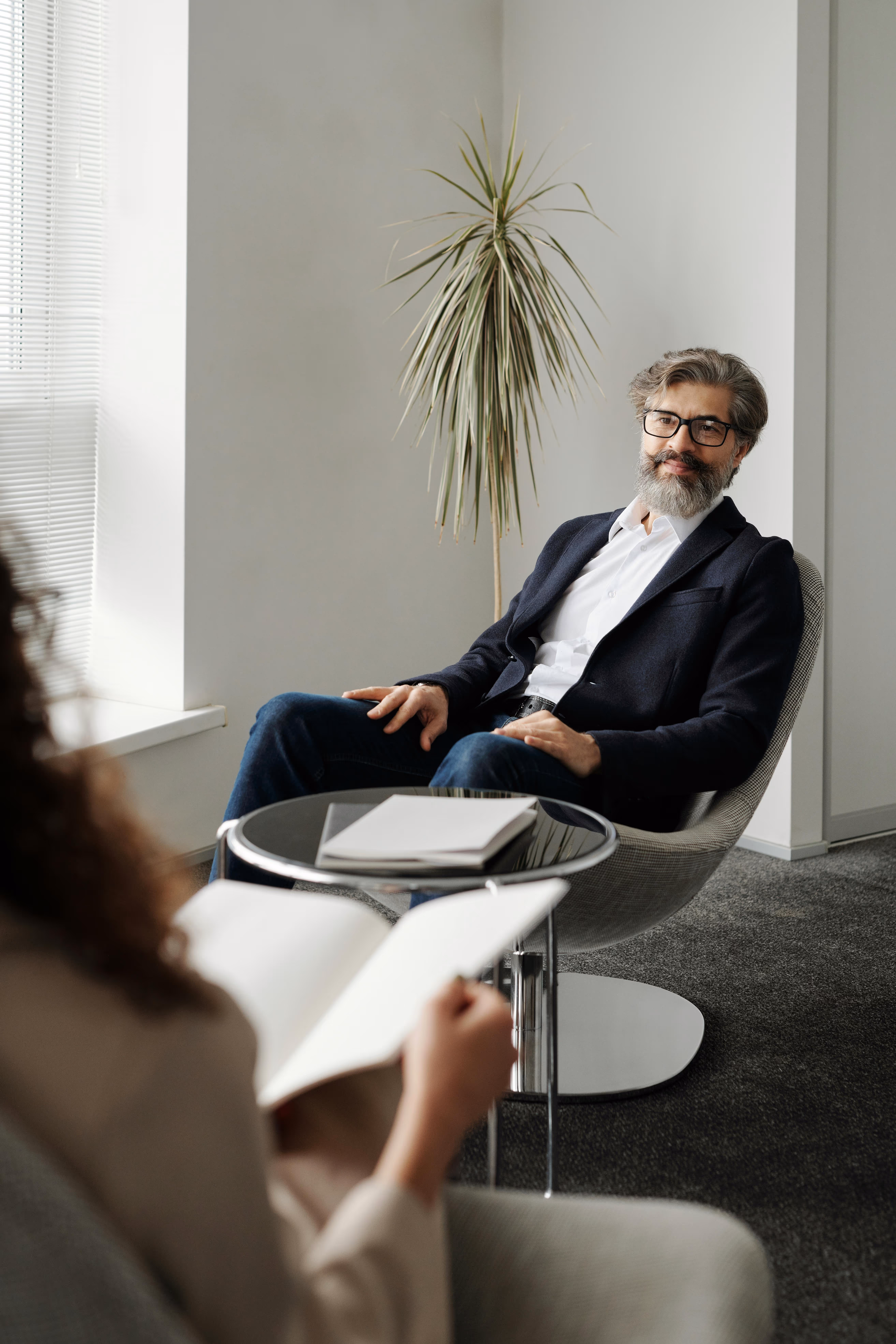 Bearded man in glasses and blazer sitting in a modern chair in an office, facing a person holding a notebook.