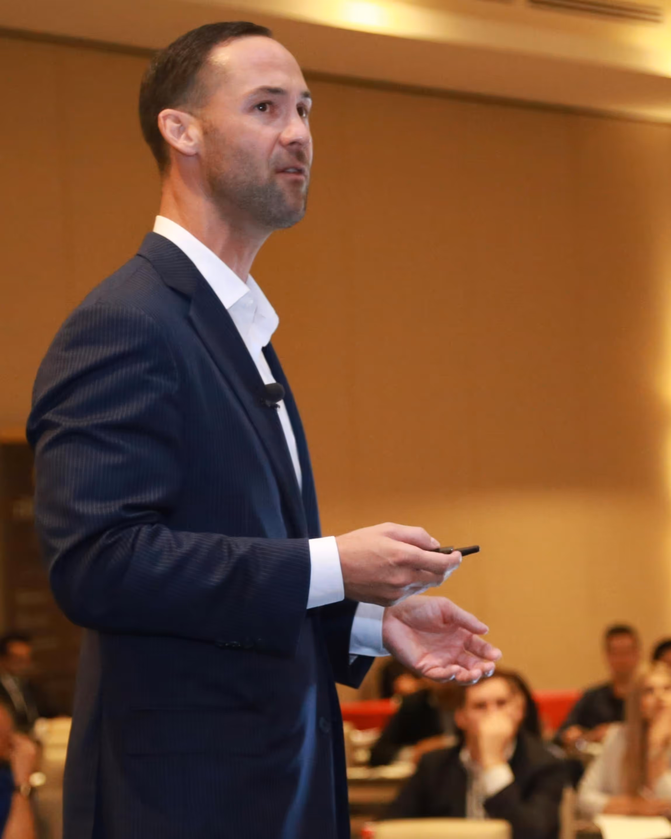 Man in a navy suit holding a remote, speaking to an audience in a conference room.