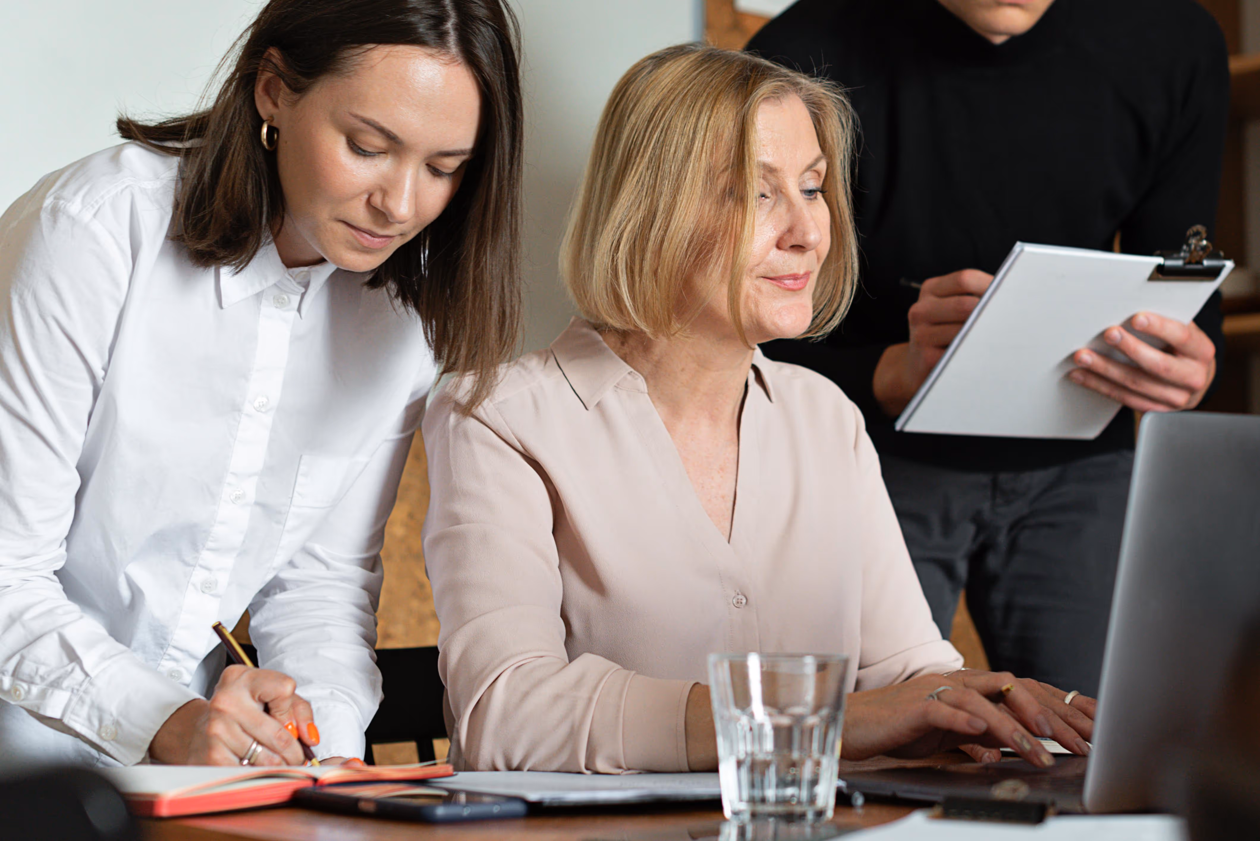 Three professionals working together with one typing on a laptop, one writing in a notebook, and one holding a clipboard.