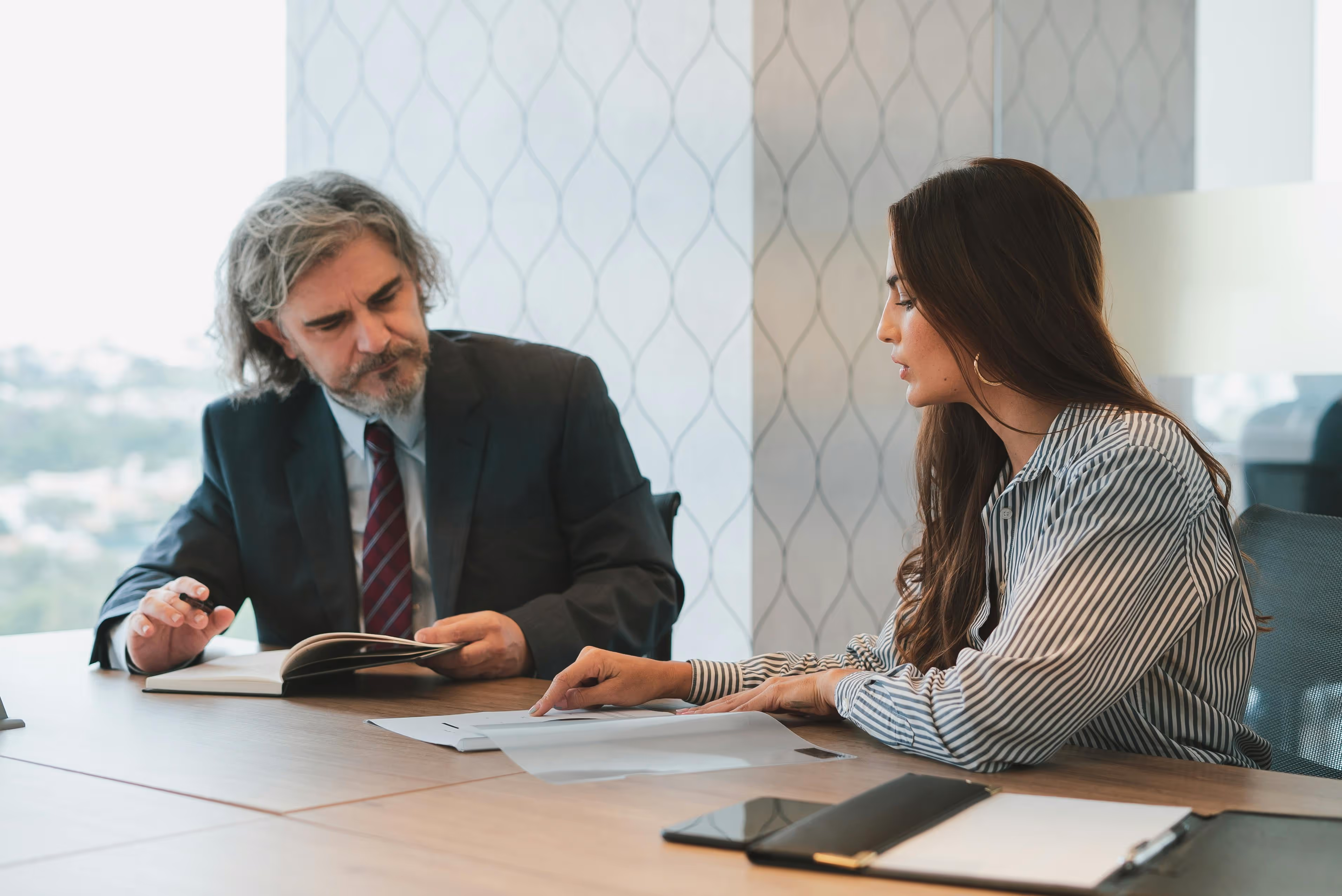 Man in suit and tie and woman in striped shirt discussing documents at a conference table in an office.