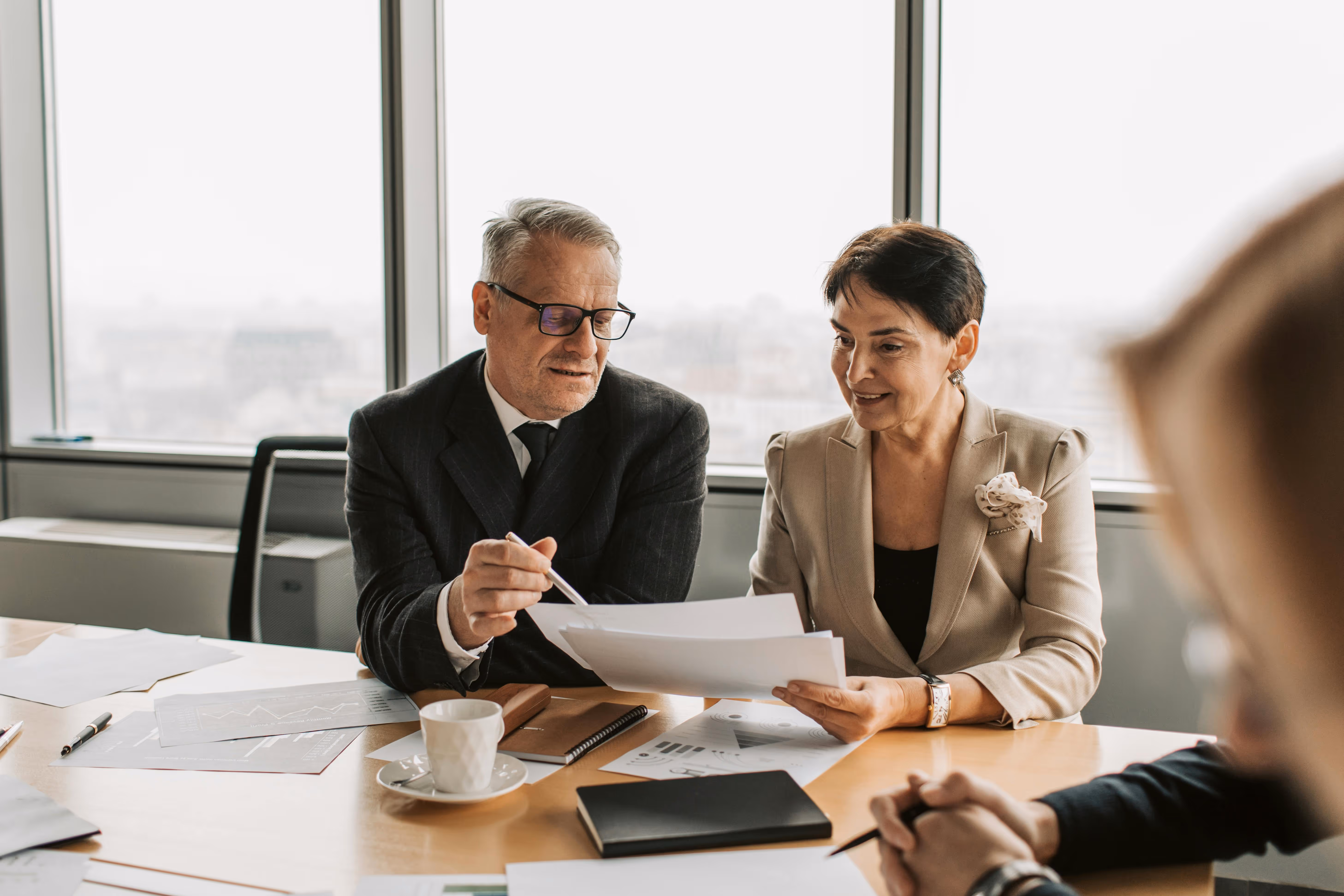 Two business professionals reviewing documents at a table in an office with large windows.