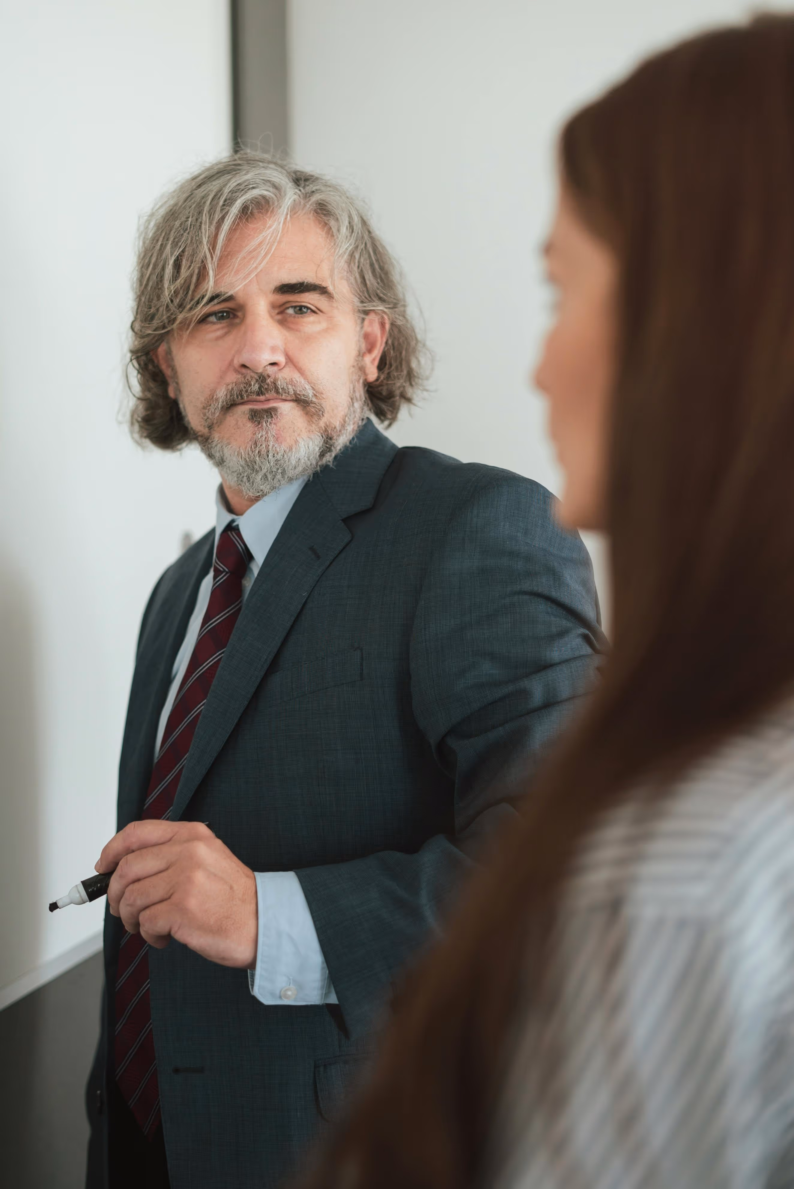Man with gray hair and beard in a suit holding a marker and looking at a woman.
