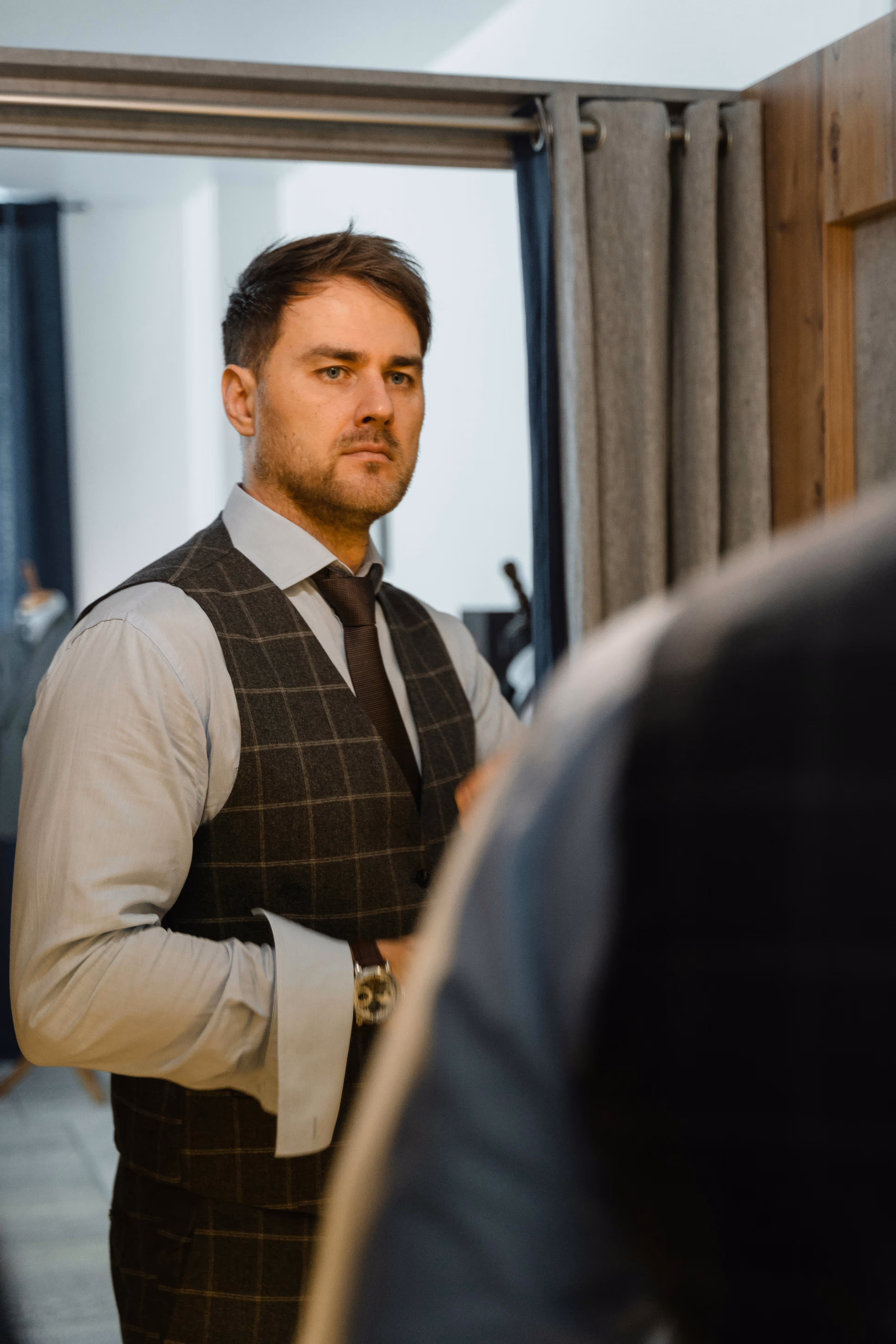 Man wearing a checkered vest and tie looking at himself in a mirror inside a room.