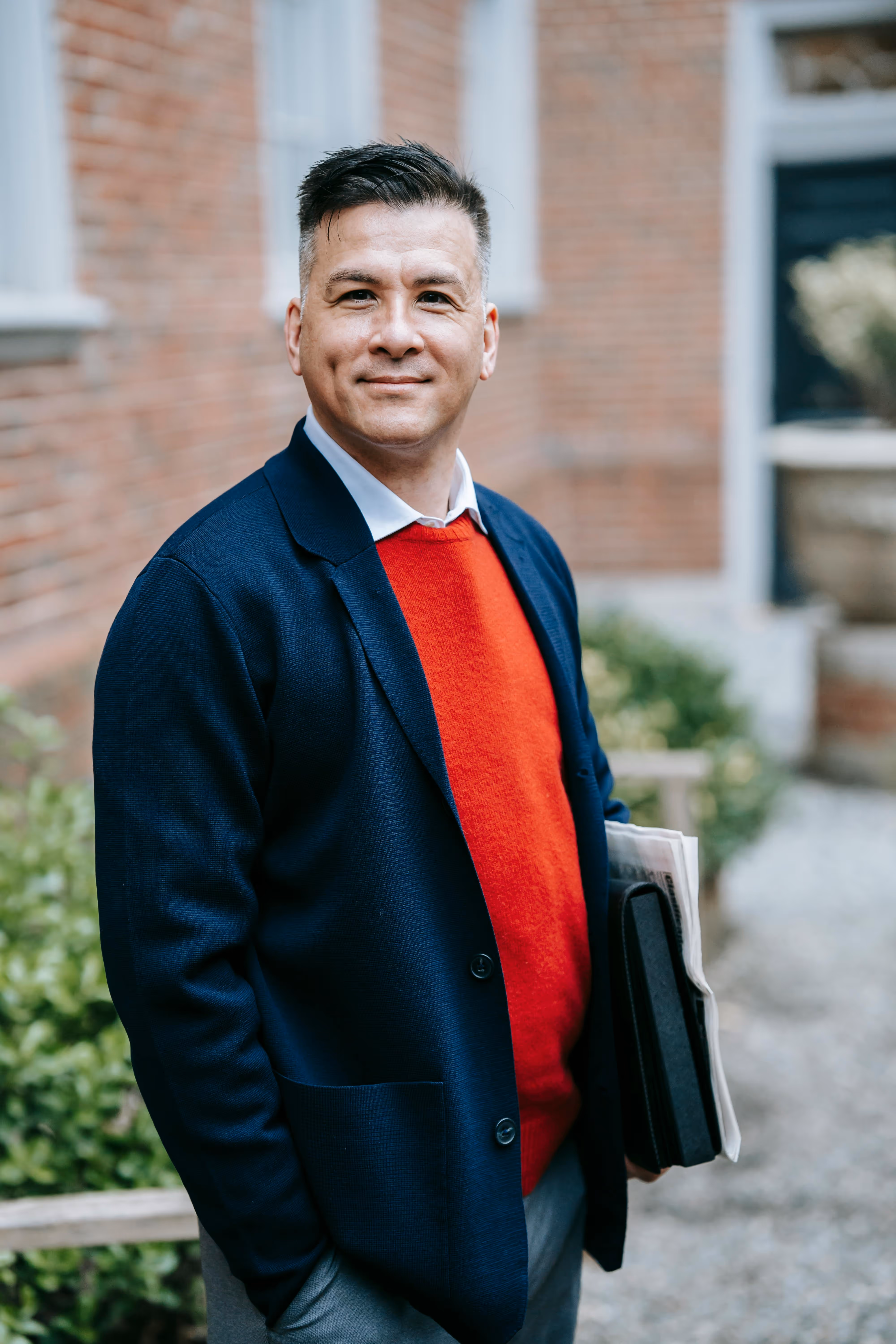 Smiling man in a navy blazer and red sweater holding folders outside a brick building.