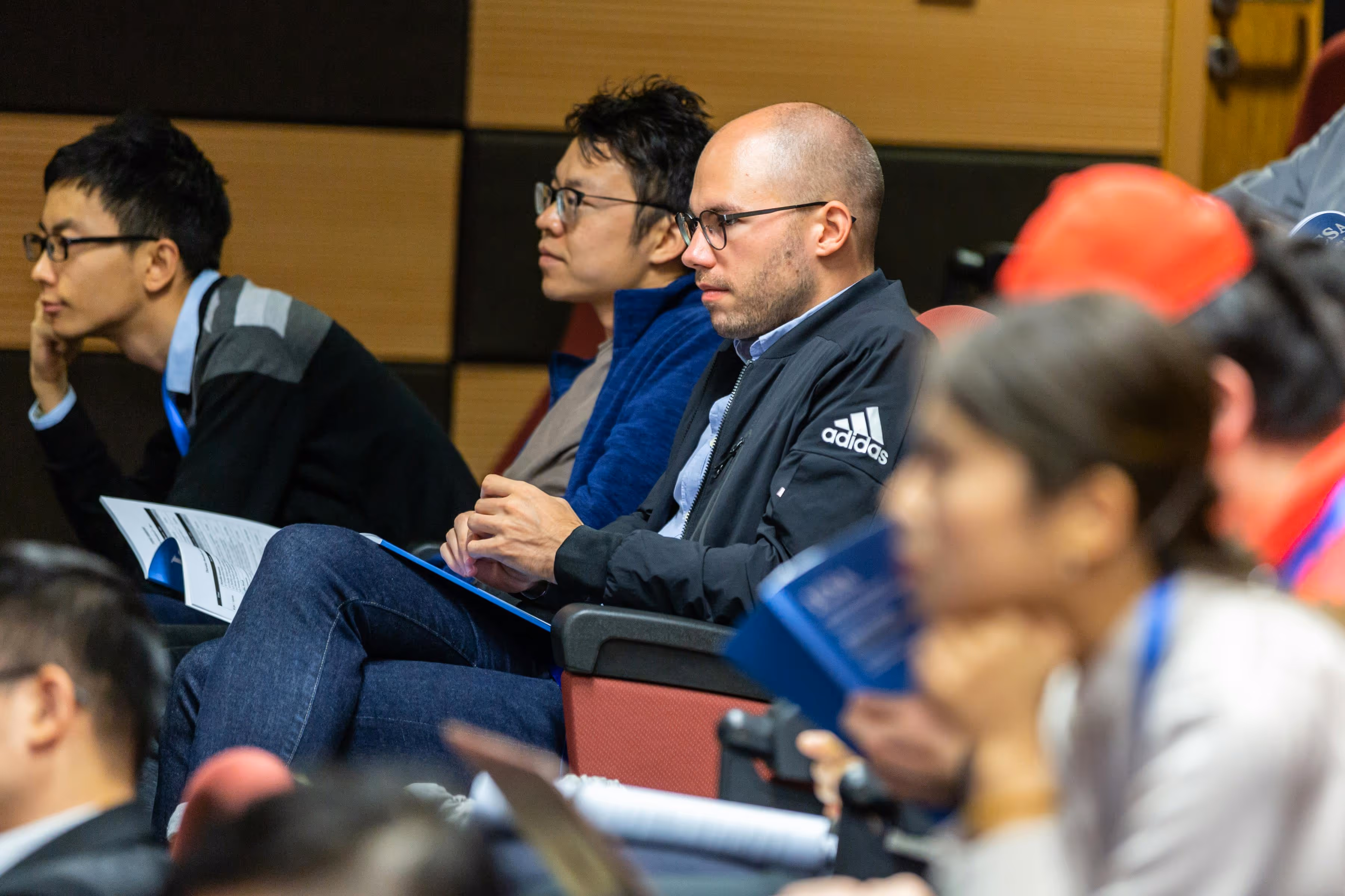 Several diverse adults seated in an auditorium attentively listening and holding conference materials.
