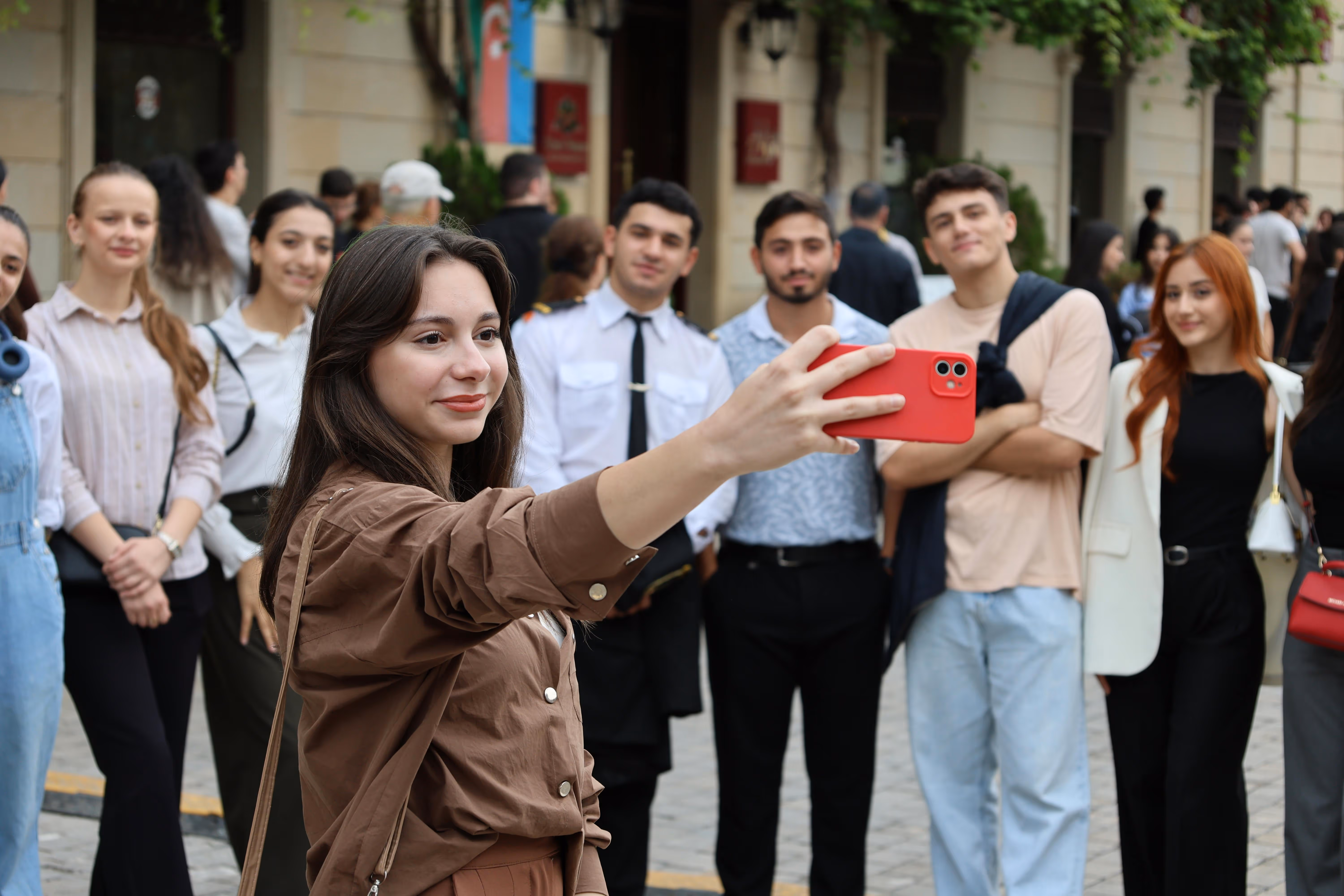 Young woman taking a selfie with a red phone, surrounded by a diverse group of smiling people outdoors.