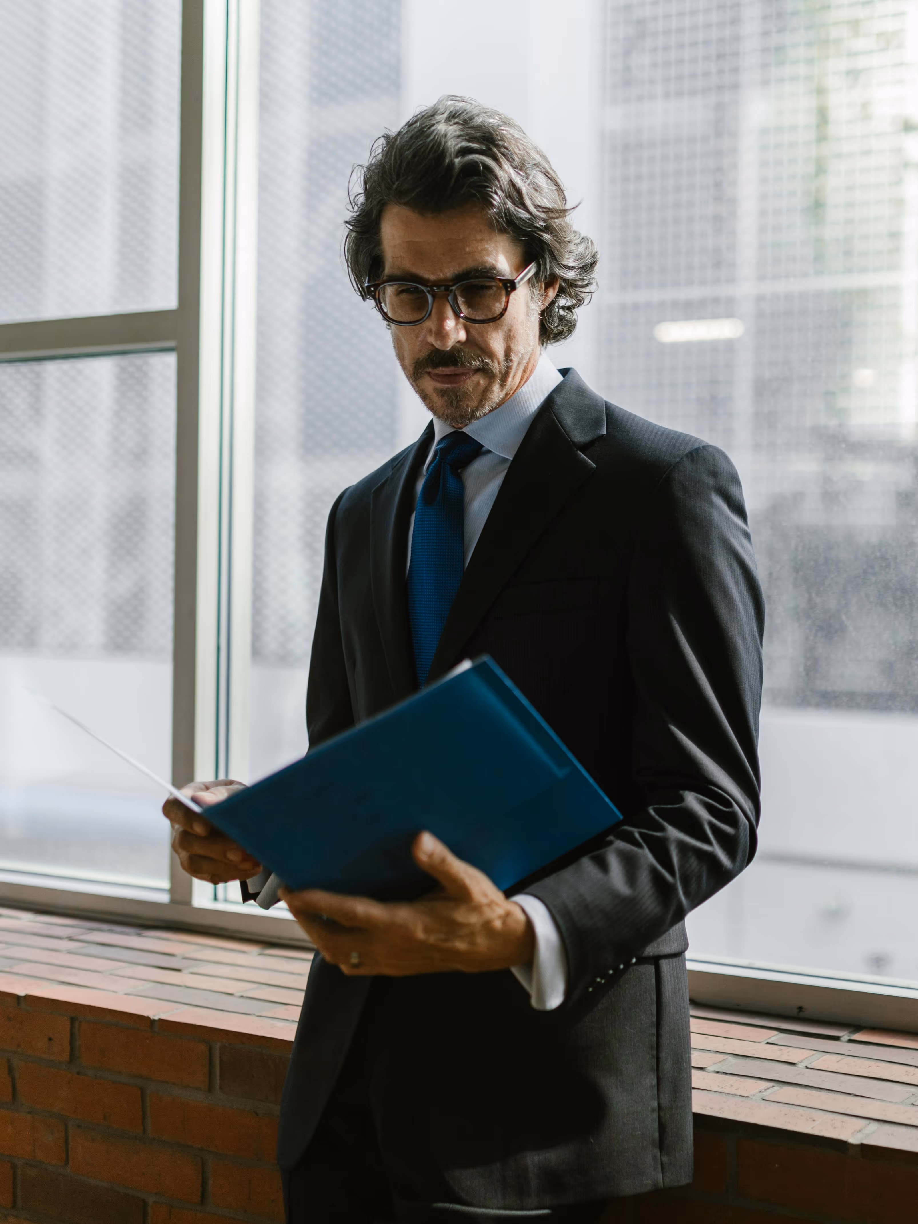 Man in a dark suit and glasses reading a blue folder by a large window with city buildings outside.