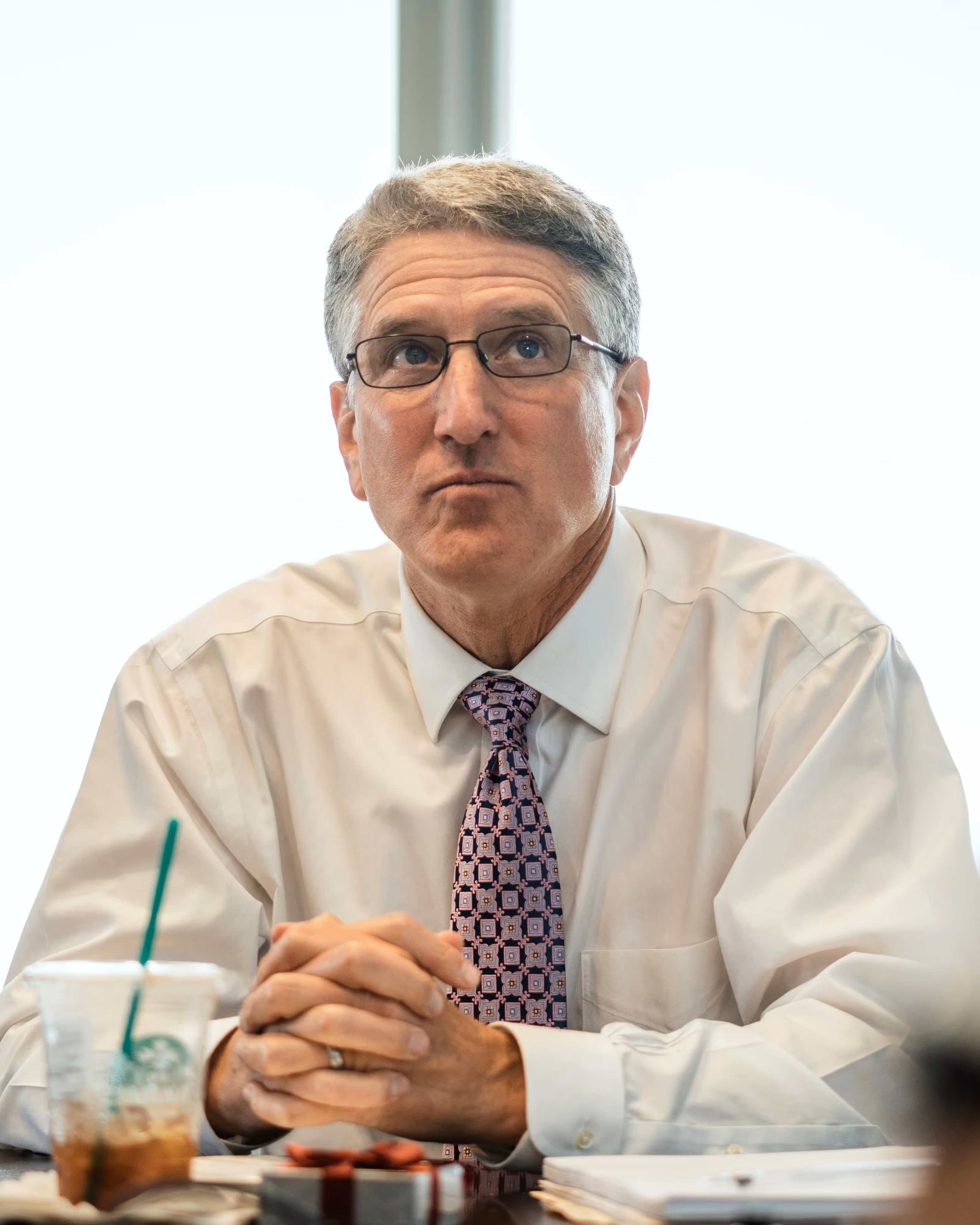 Middle-aged man wearing glasses, a white shirt, and patterned tie sitting at a table with folded hands, looking thoughtfully upward.