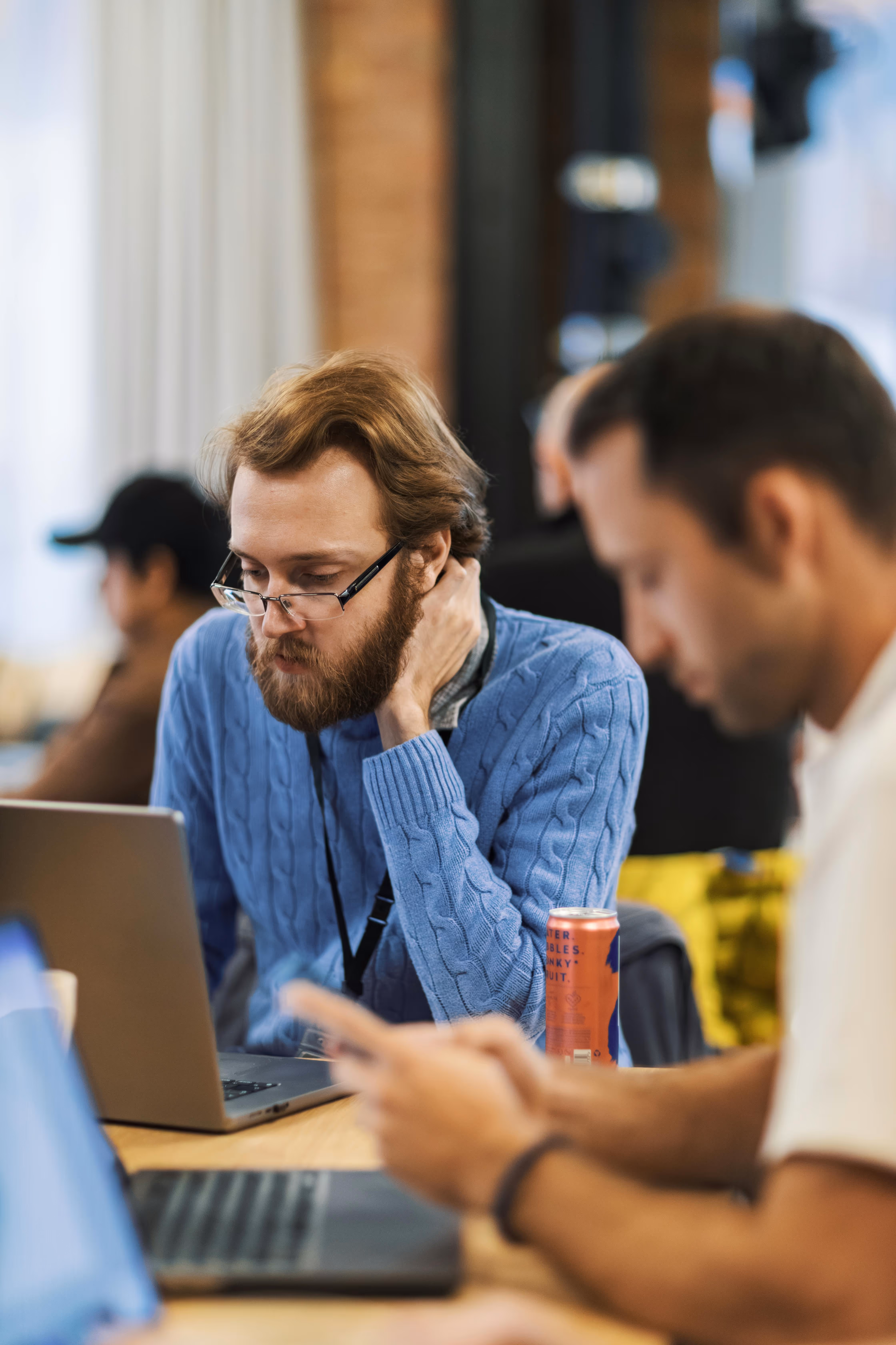 Bearded man in glasses and blue sweater working on a laptop in a shared workspace.