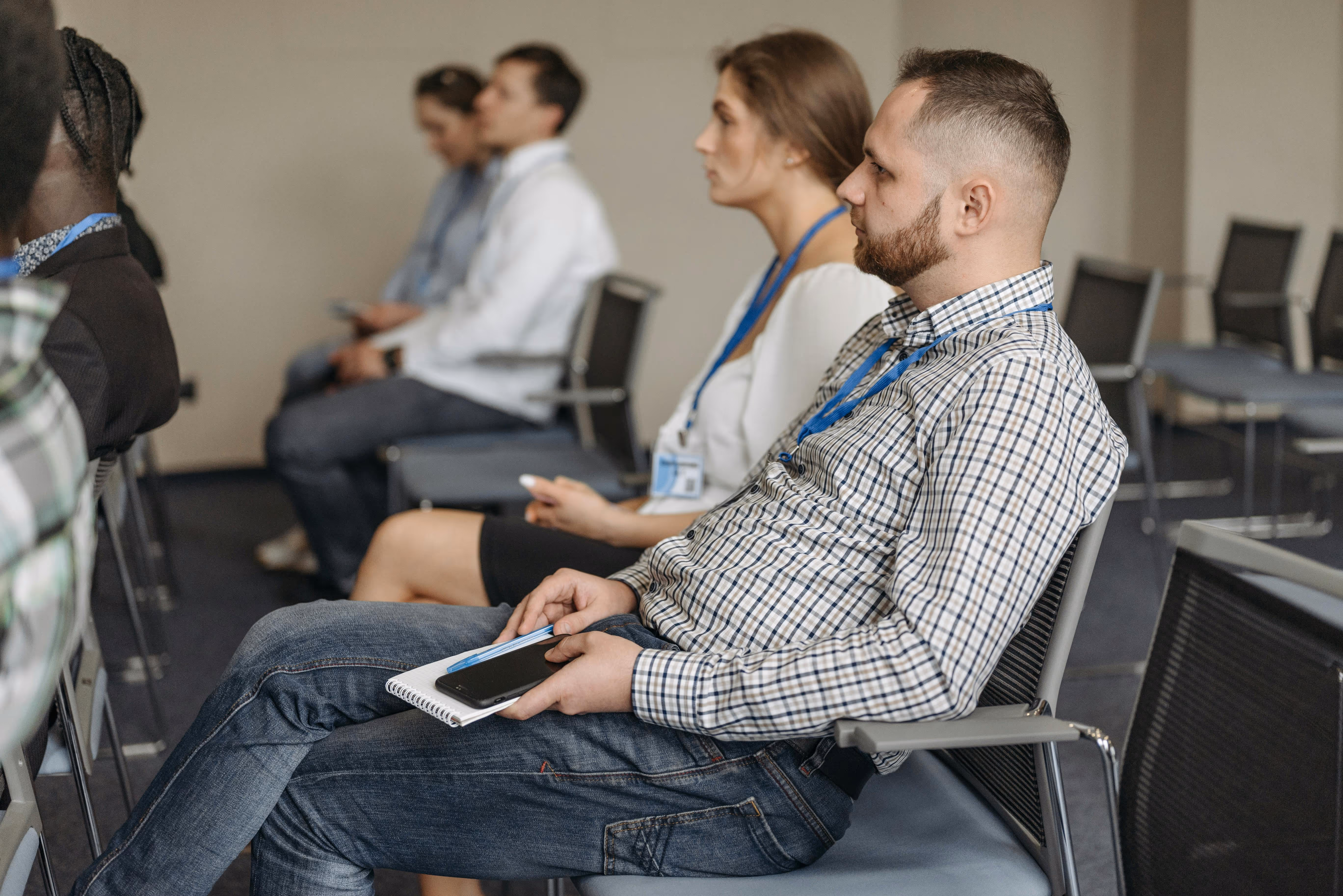 Audience members with conference badges seated in a room, one man in a plaid shirt holding a notebook and phone.