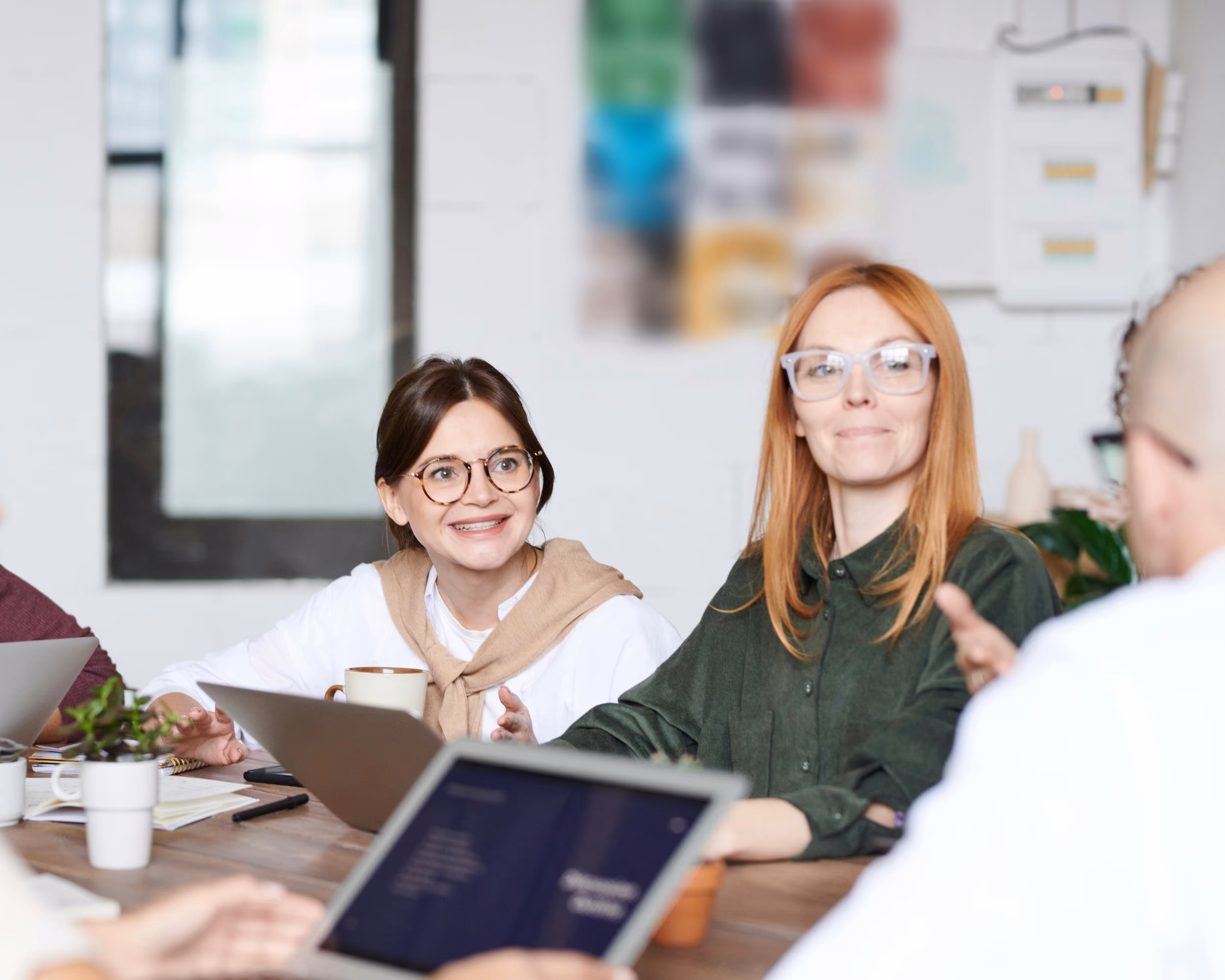 Two women wearing glasses engaged in conversation at a meeting table with laptops and coffee mugs.