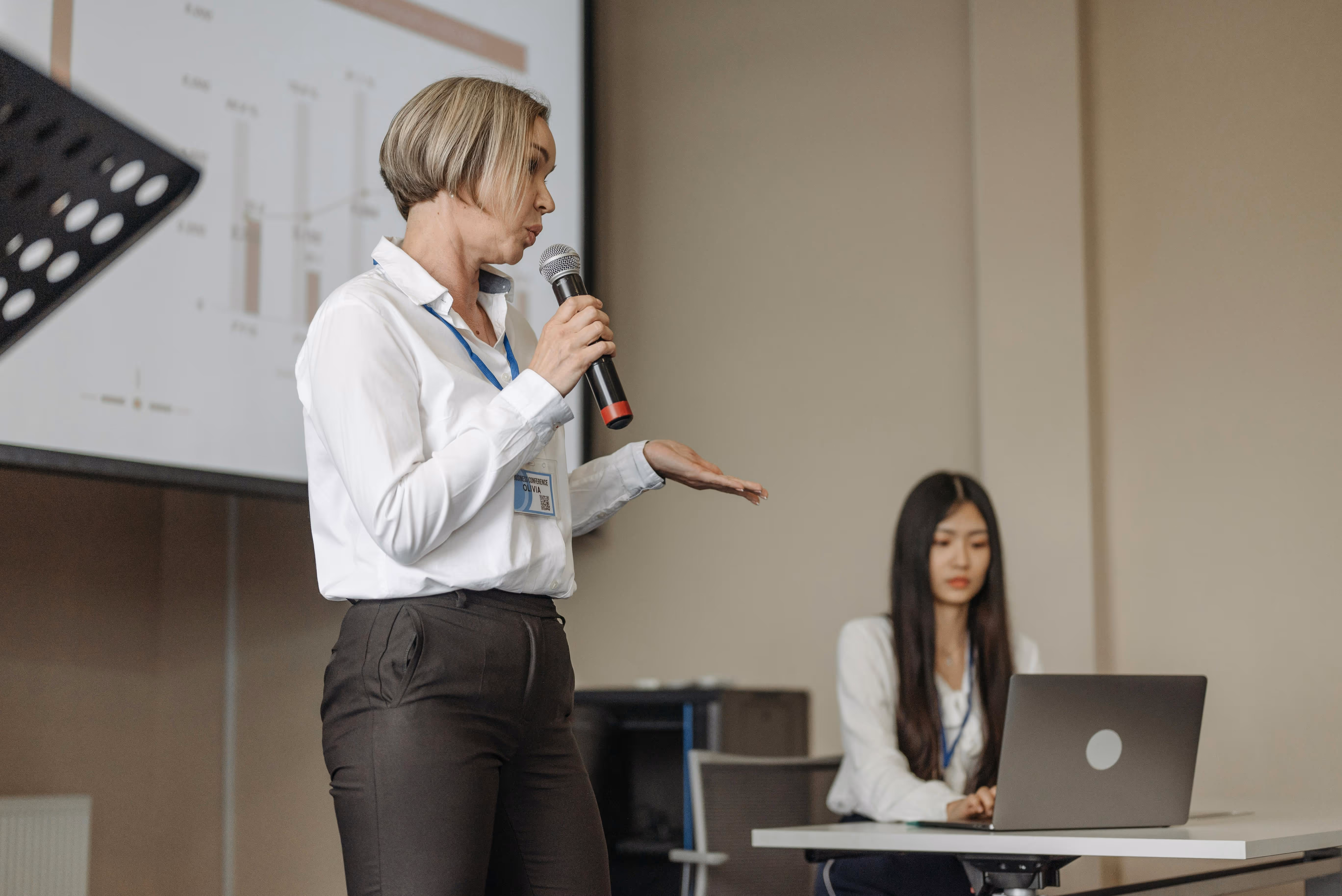 Woman speaking into a microphone during a presentation with a seated woman working on a laptop in the background.