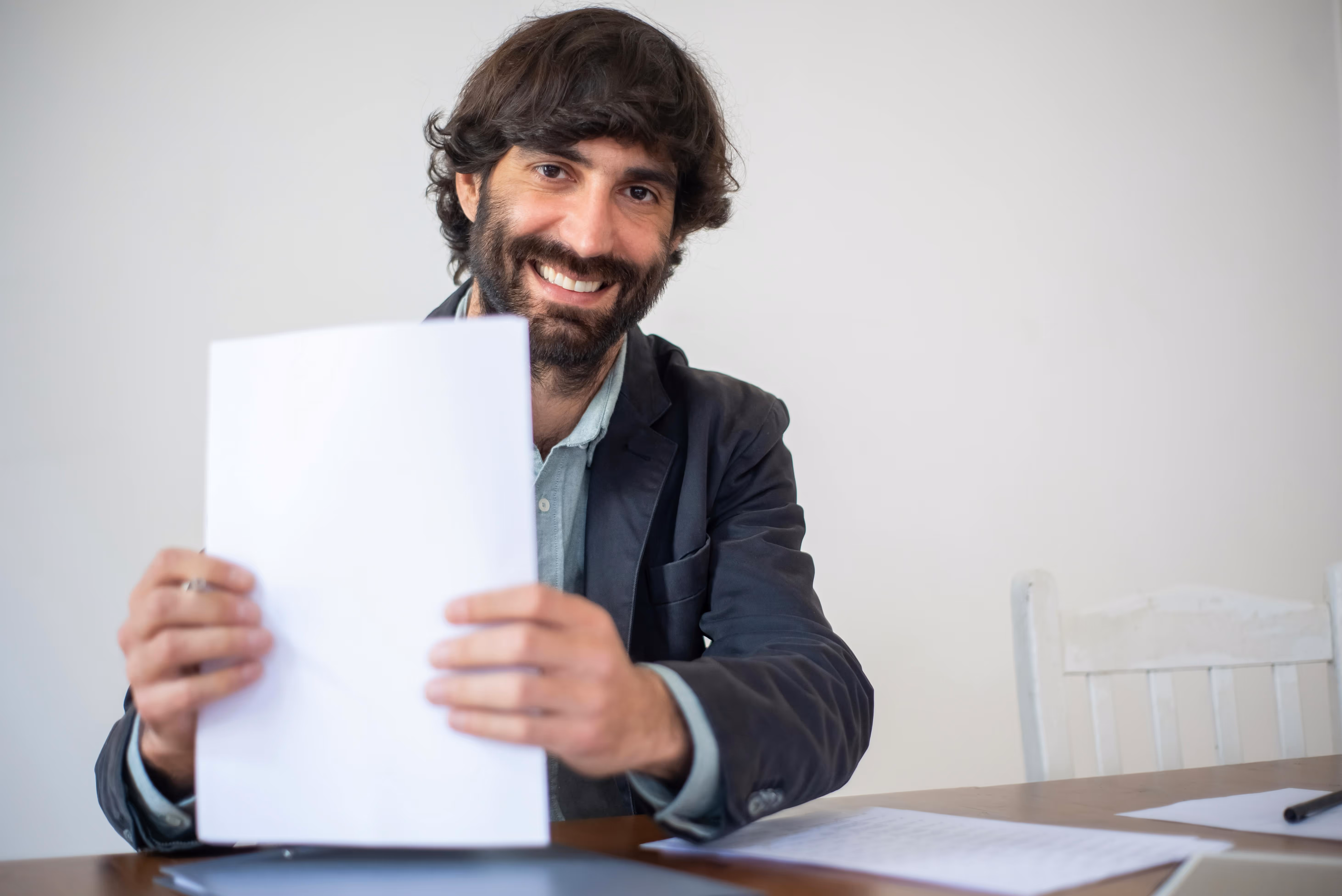 Smiling man with dark hair and beard holding up a blank sheet of paper at a desk.