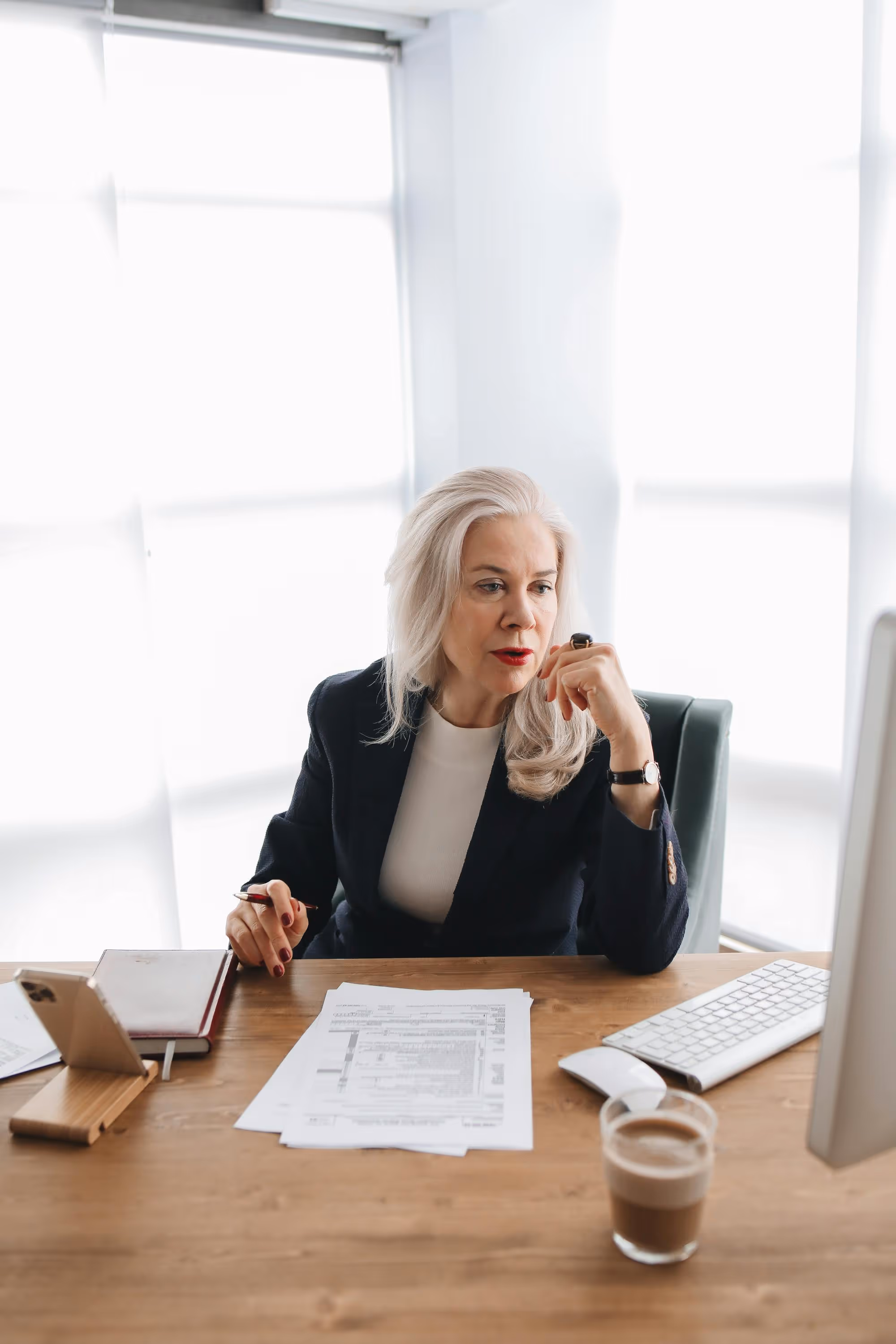 Mature woman with white hair in a black blazer sitting at a desk reviewing documents with a pen in hand.