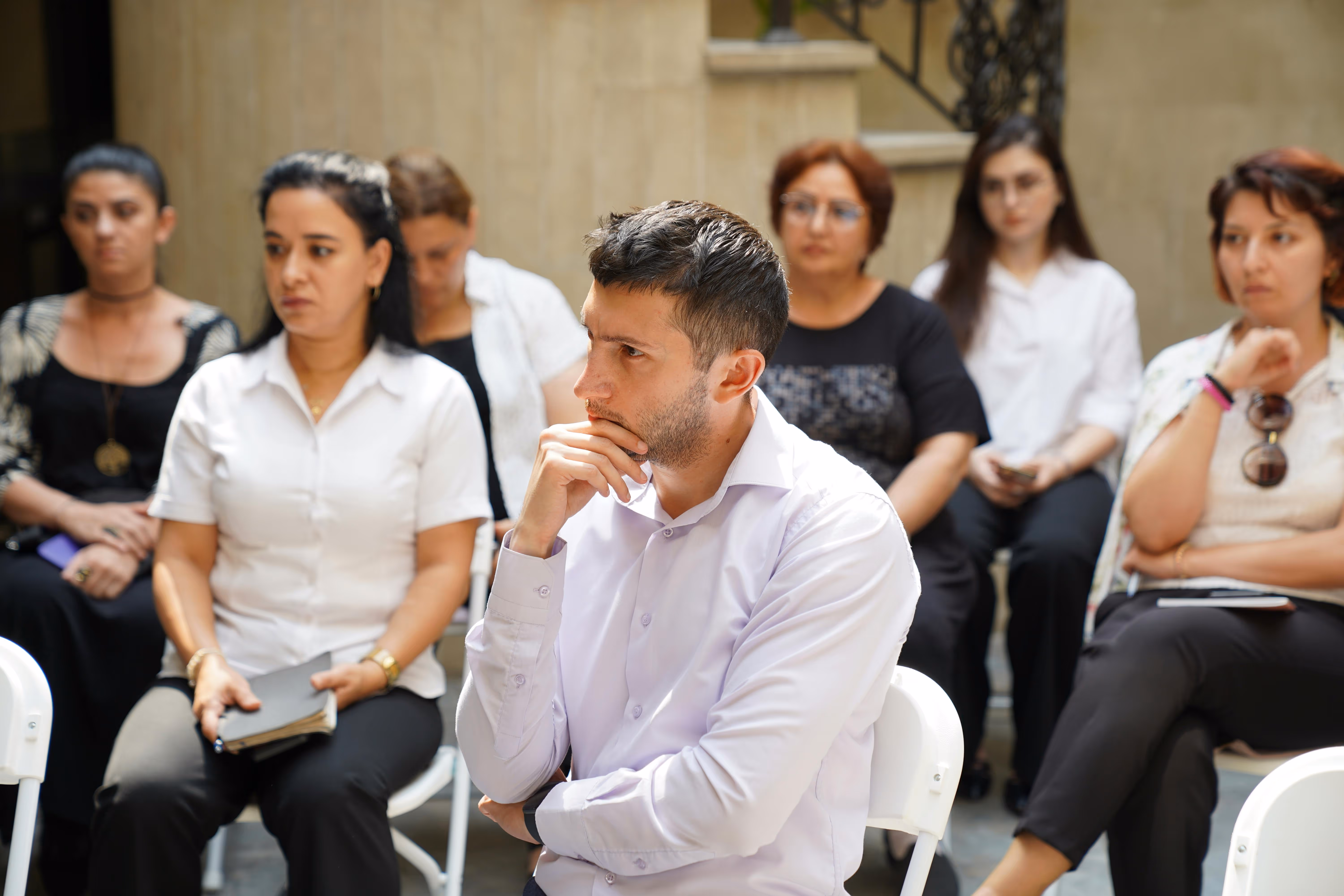Man in a white shirt sitting with hand on chin, attentively listening in a group of seated people.