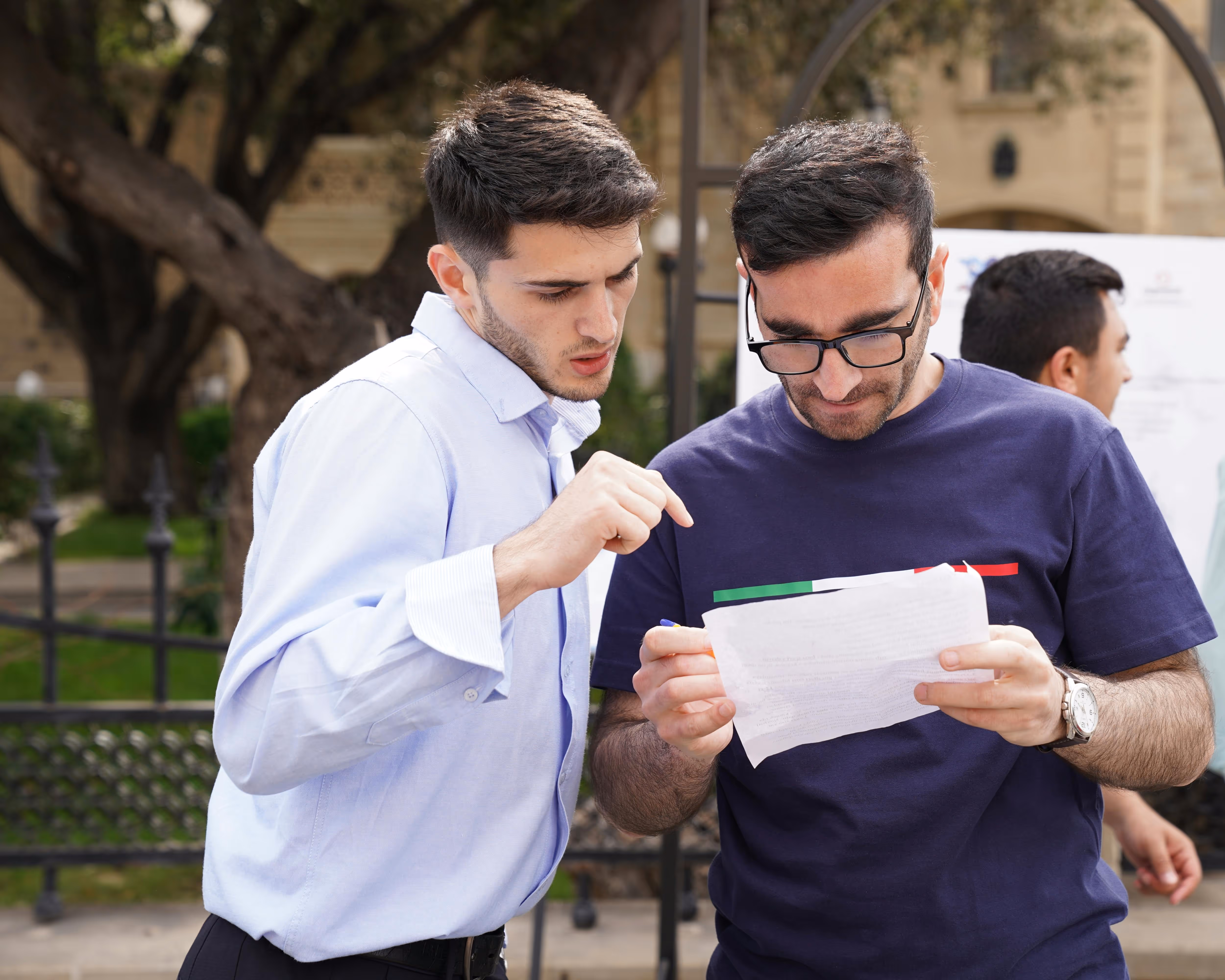 Two men outdoors closely reading and discussing a document, one wearing a light blue shirt and the other wearing glasses and a navy t-shirt.