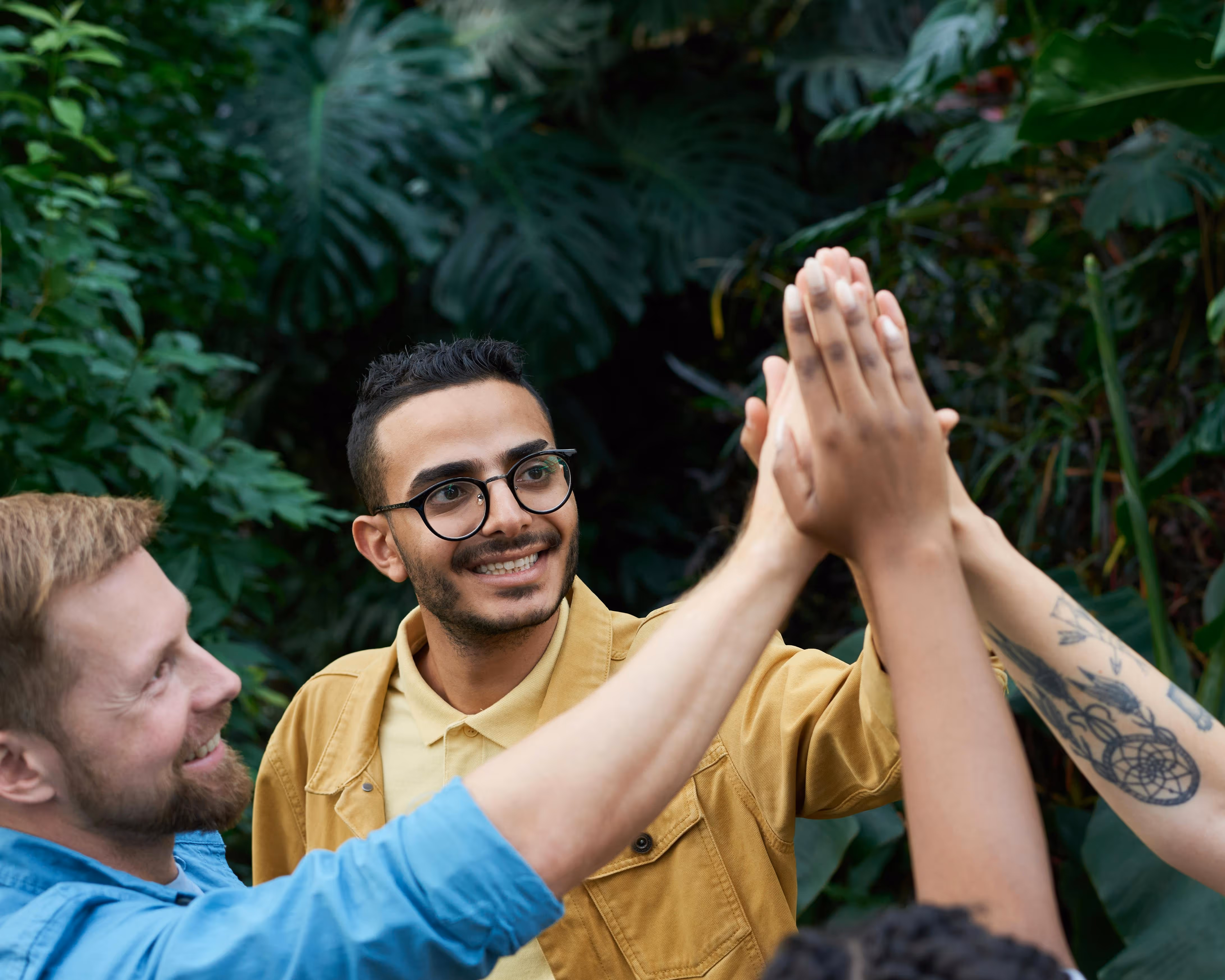A diverse group of people smiling and giving a high-five outdoors with lush green plants in the background.