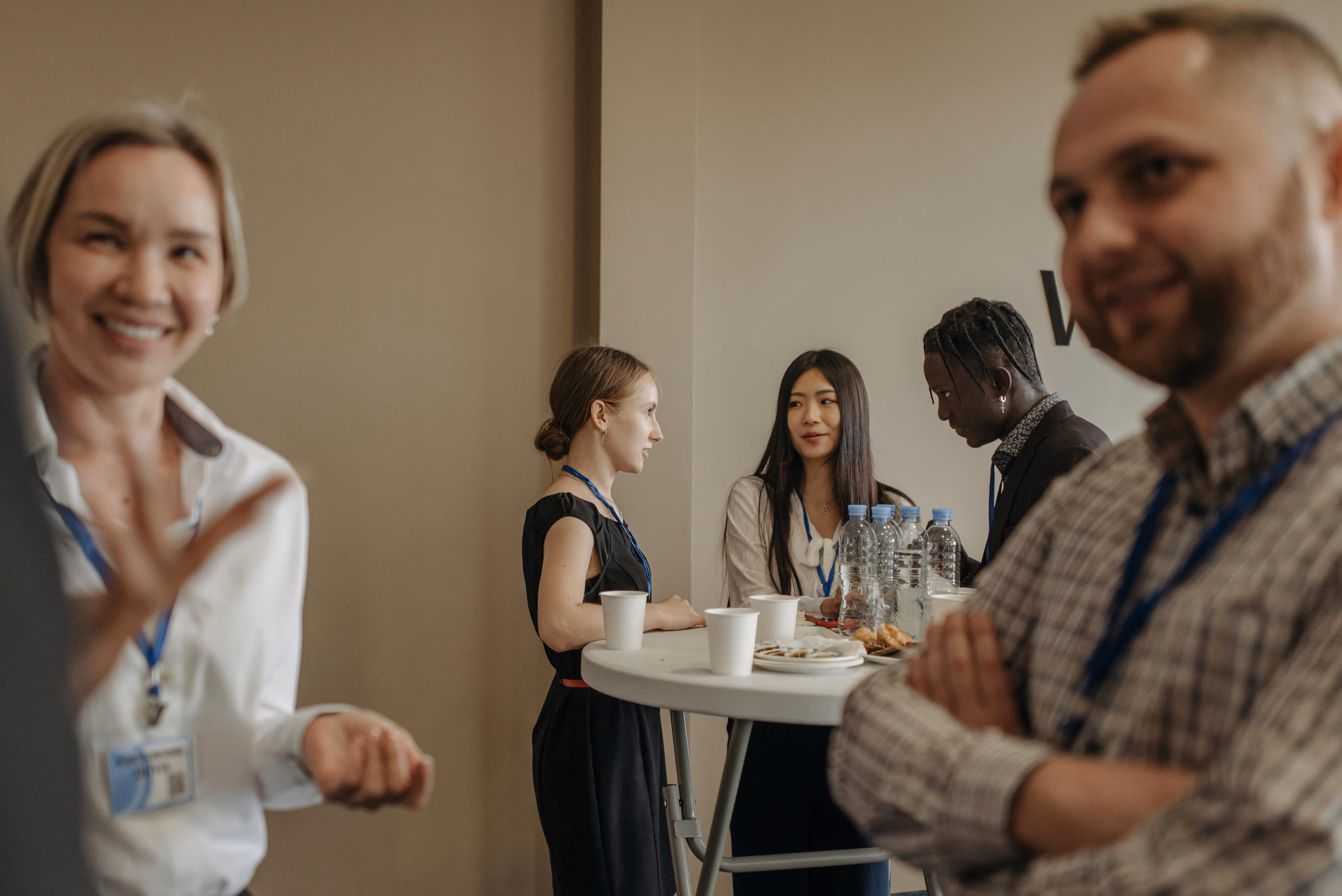 Five diverse people wearing conference badges standing and conversing around a tall table with water bottles and snacks.