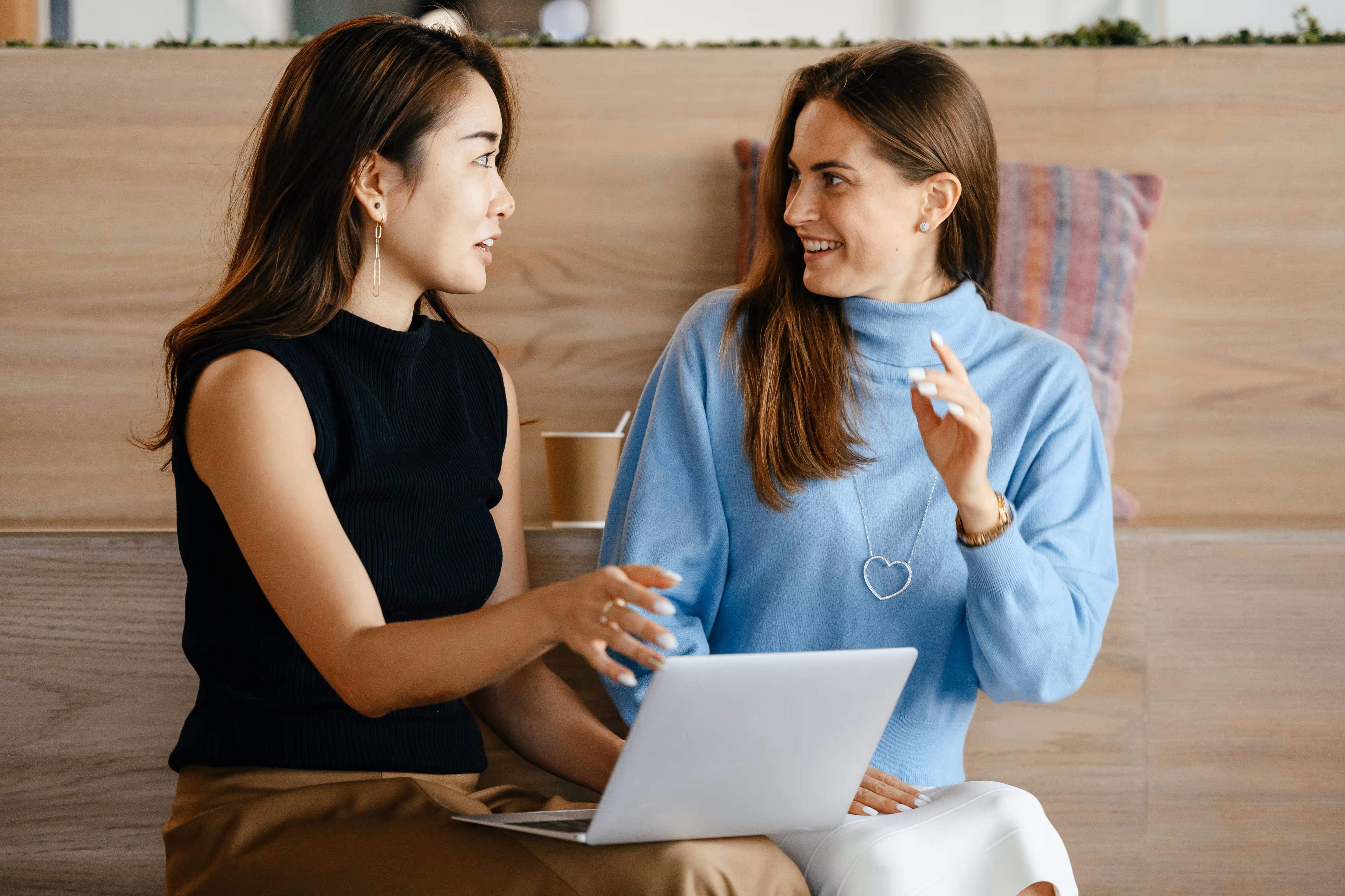 Two women sitting and smiling at each other while using a laptop in a casual indoor setting.