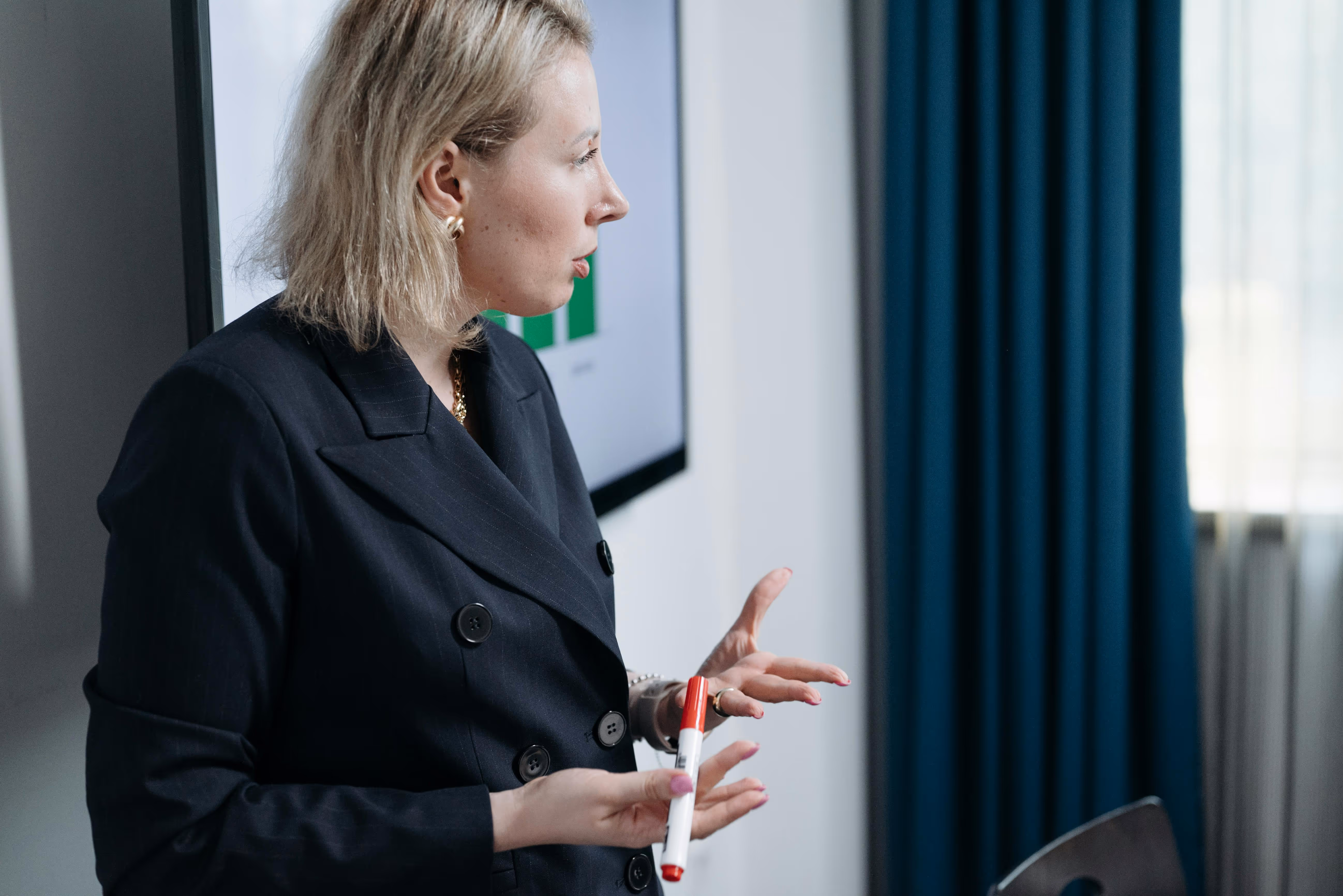 Woman in a black blazer holding a red and white marker, speaking with a presentation screen in the background.
