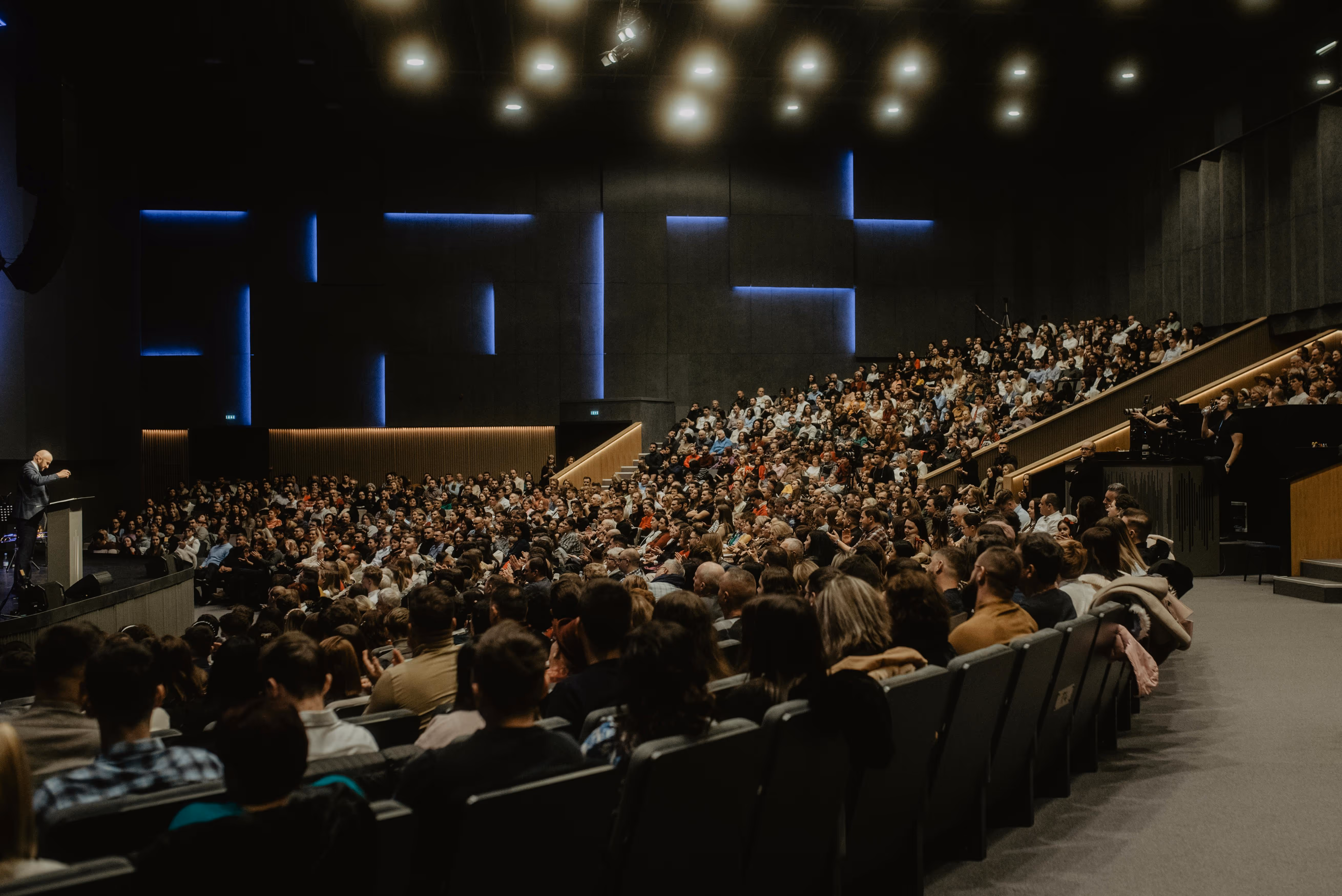 Large audience seated in a modern auditorium listening to a speaker at a podium.