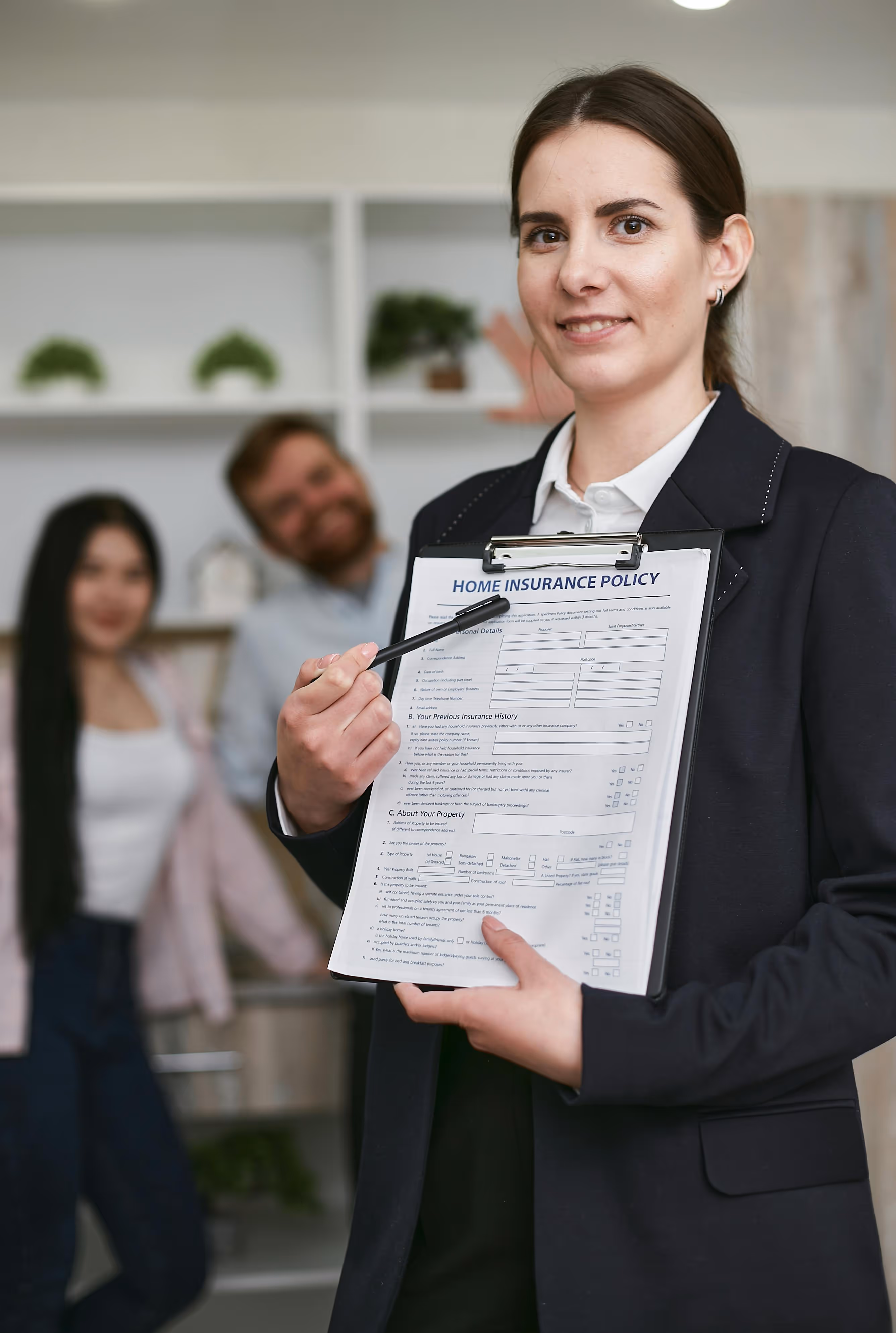 Woman in business attire holding a clipboard with a home insurance policy form, pointing at the document with a pen, with a smiling couple blurred in the background.