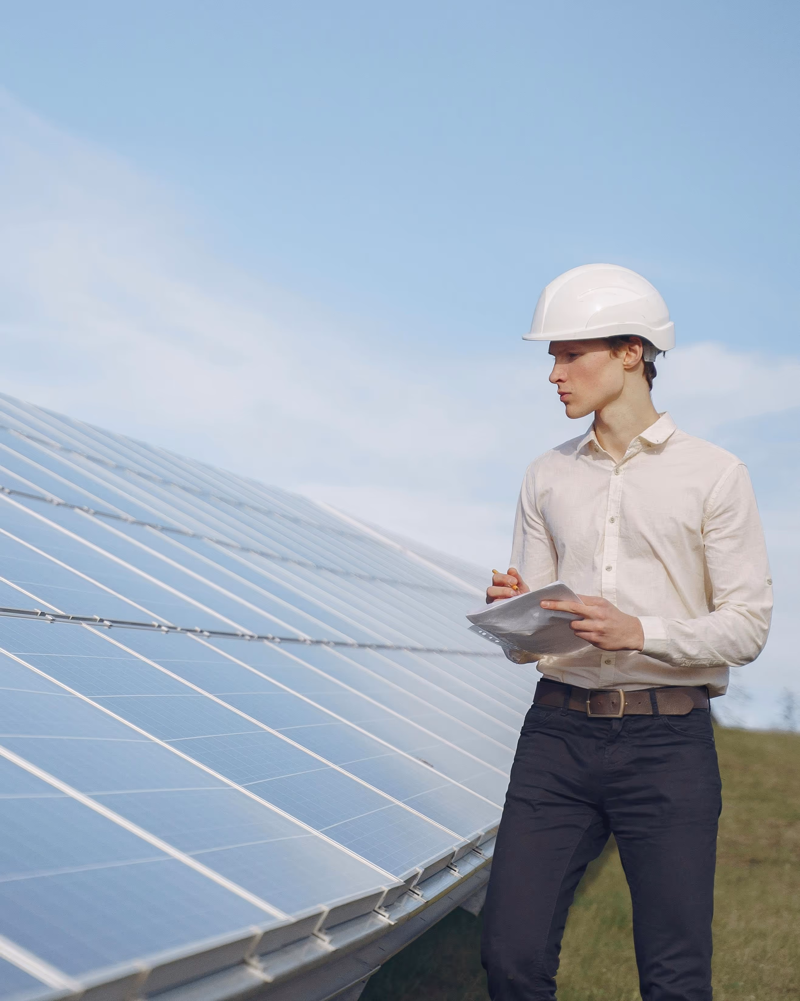 Man in a white hard hat and light shirt inspecting solar panels outdoors while holding a clipboard and pencil.