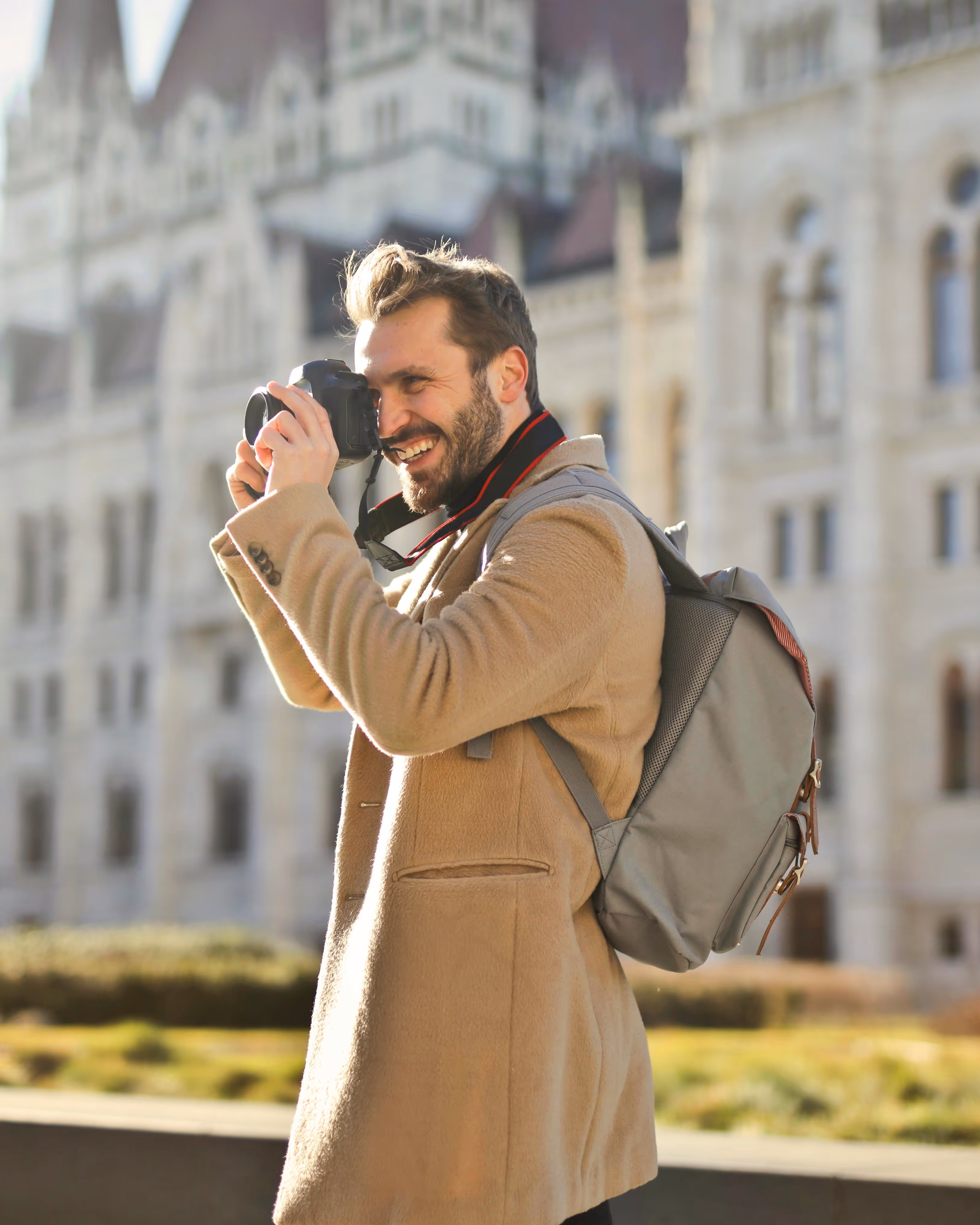 Smiling man in a beige coat with a gray backpack taking a photo with a camera in front of a historic building.