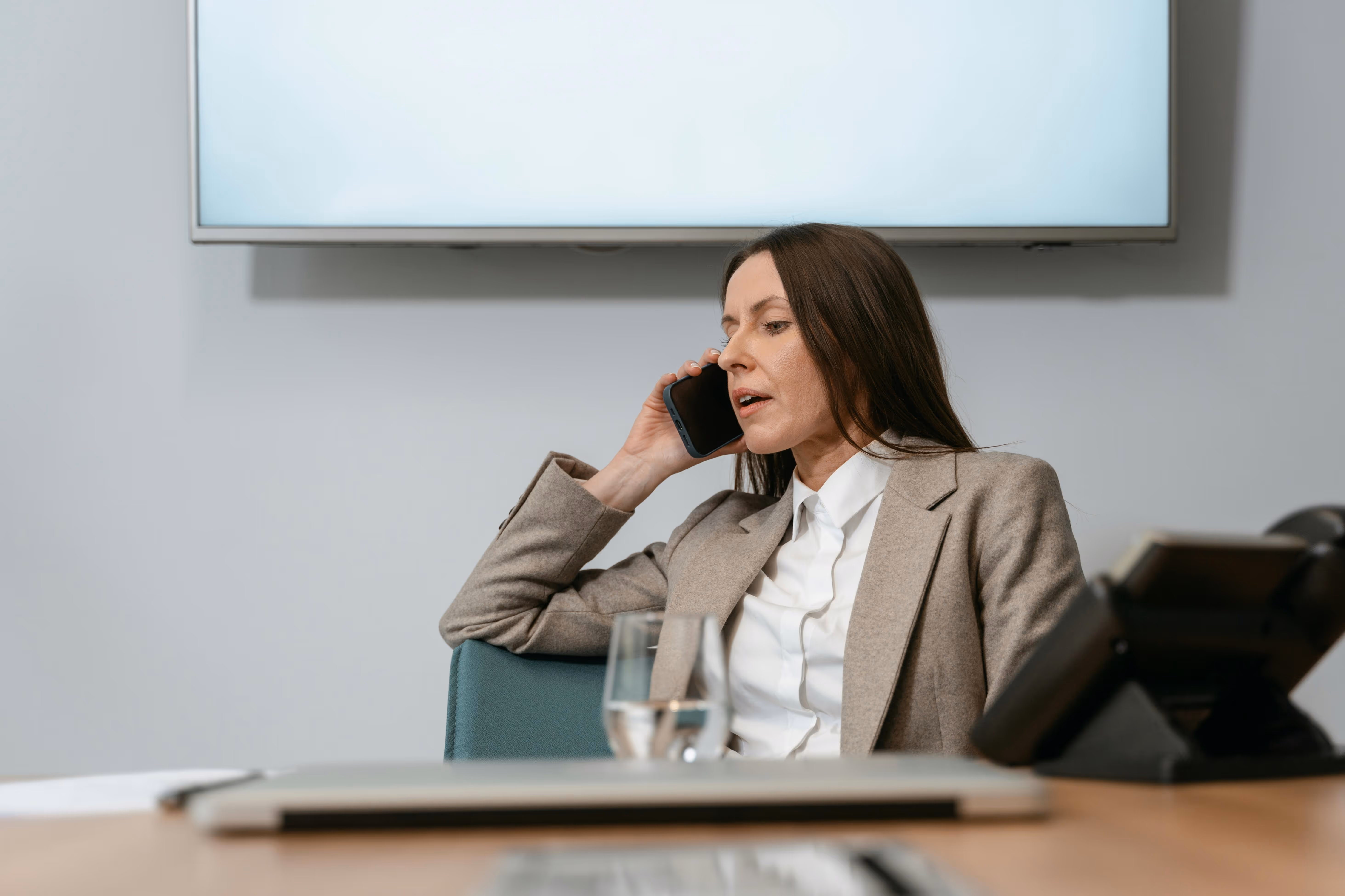 Businesswoman in beige blazer talking on a smartphone while seated at a conference table with a glass of water and office phone nearby.