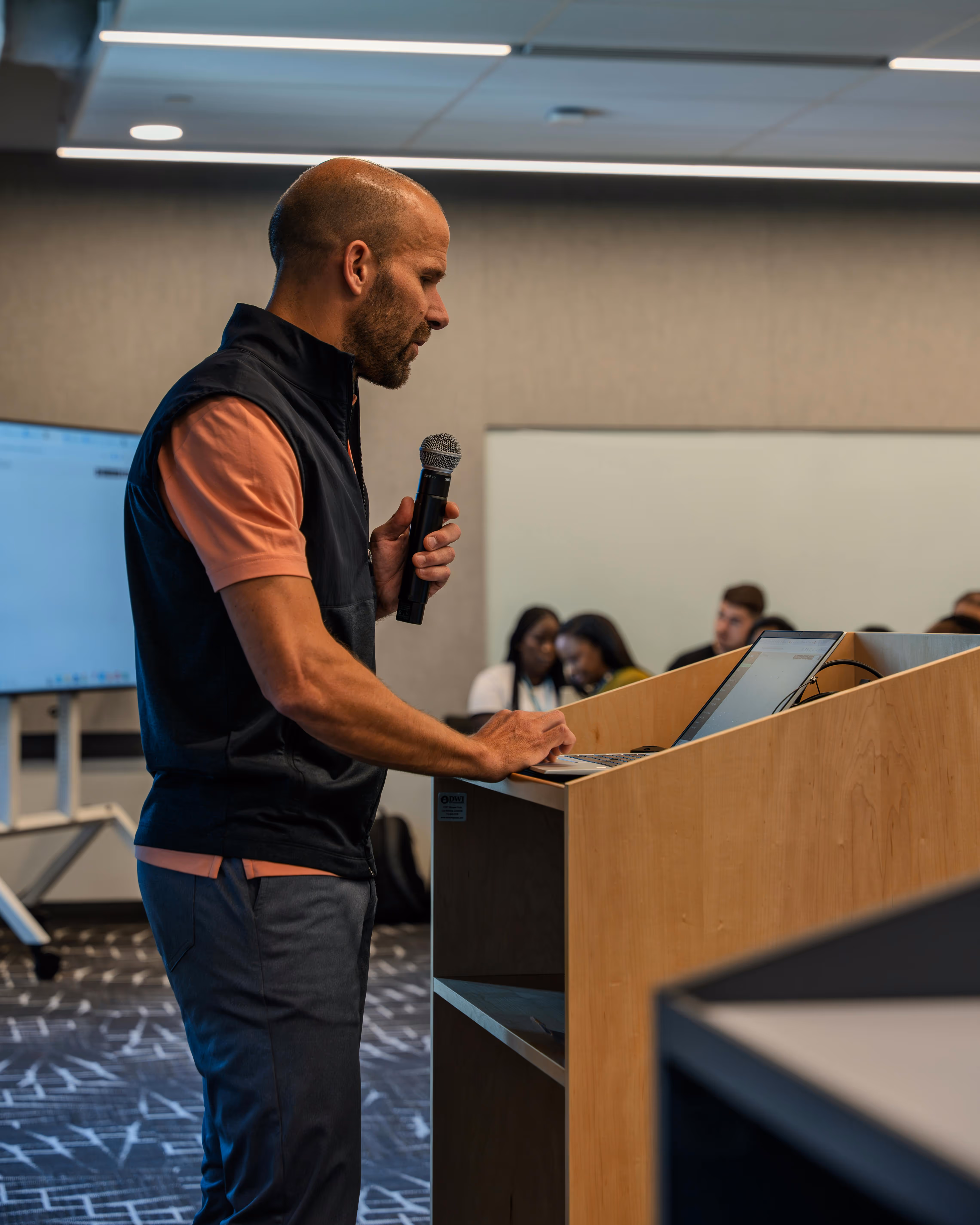 Man holding a microphone and typing on a laptop at a wooden podium in a conference room with audience in the background.