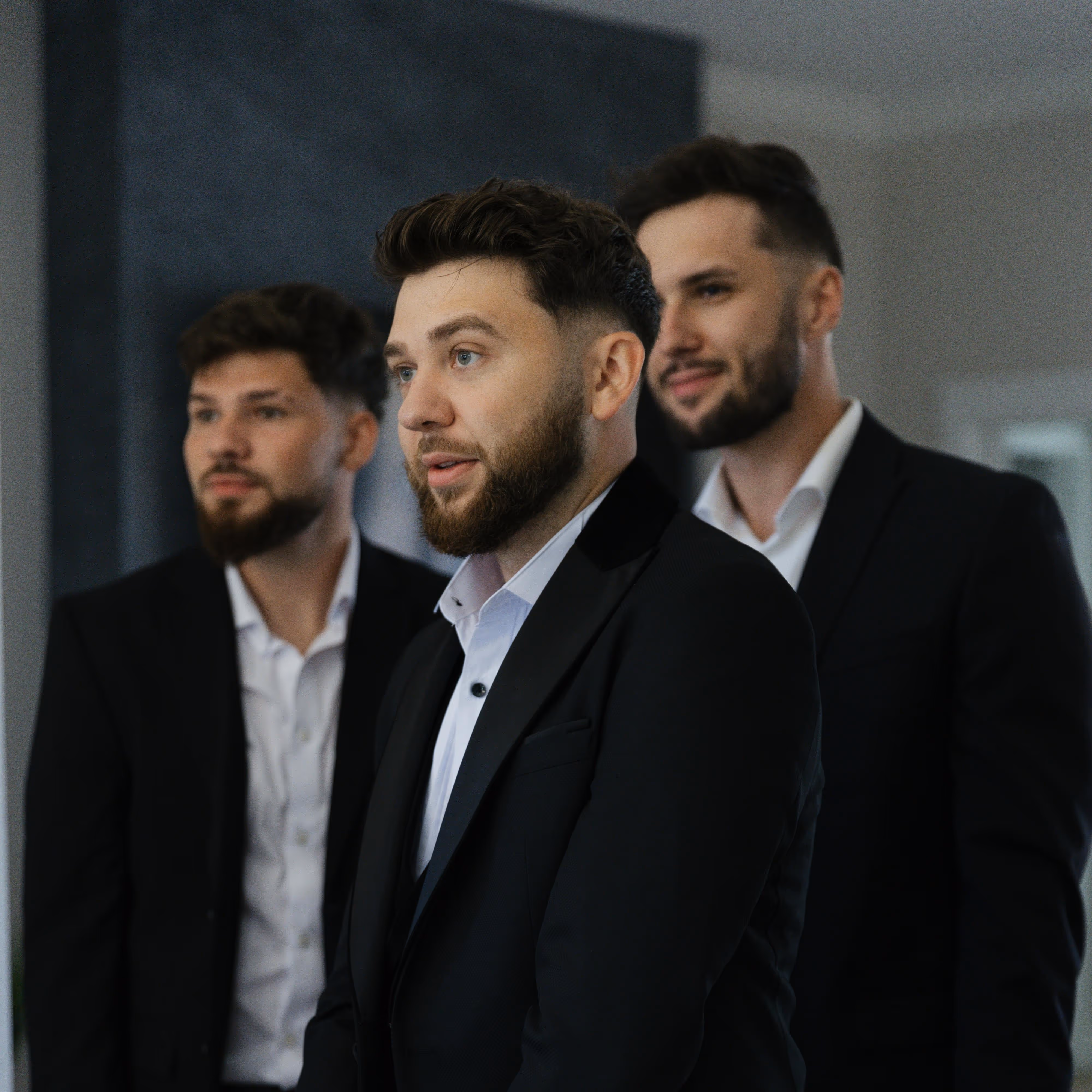 Three young men with beards wearing black suits and white shirts standing indoors, looking slightly to the right.