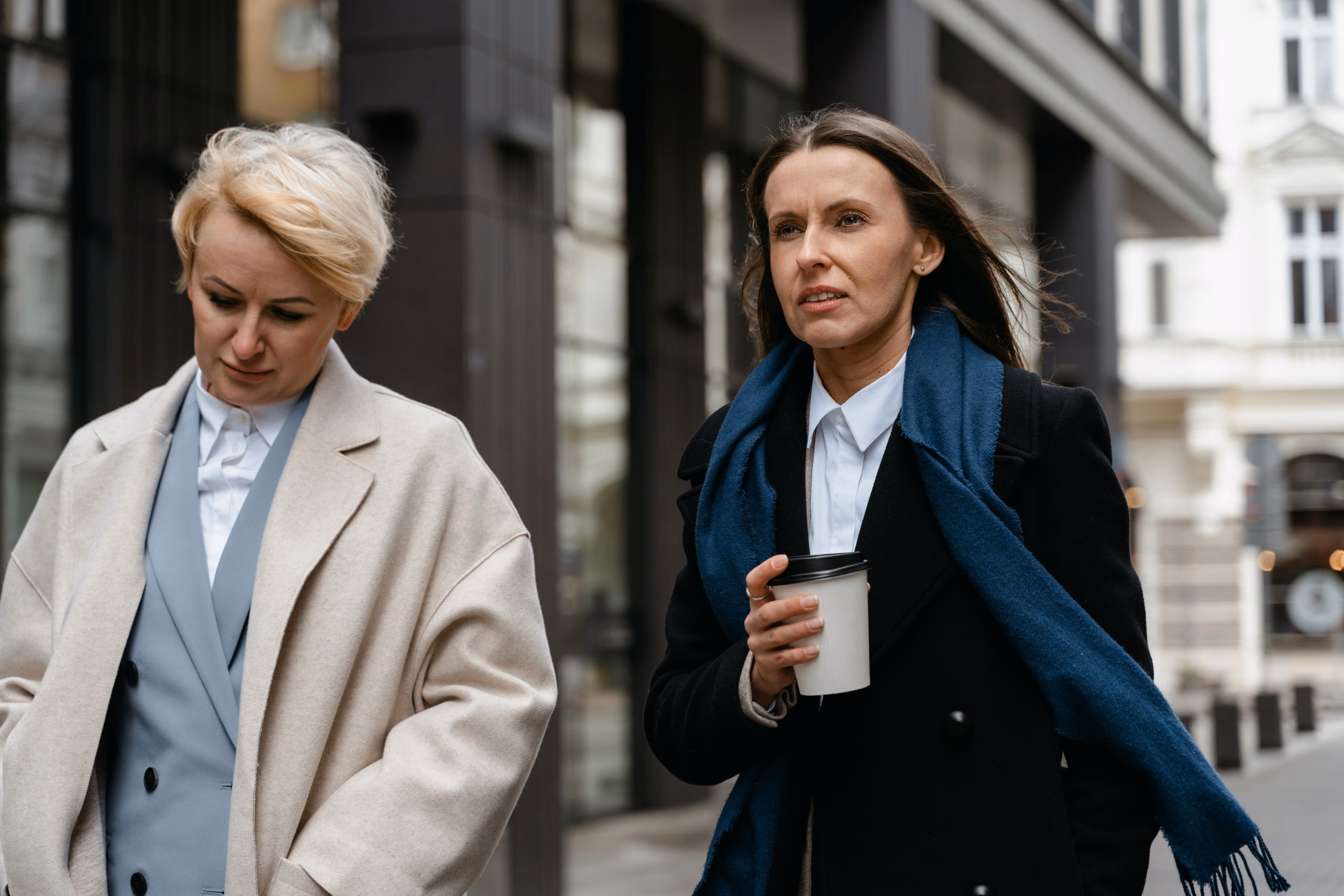 Two women walking on a city street, one holding a takeout coffee cup and wearing a black coat with a blue scarf.