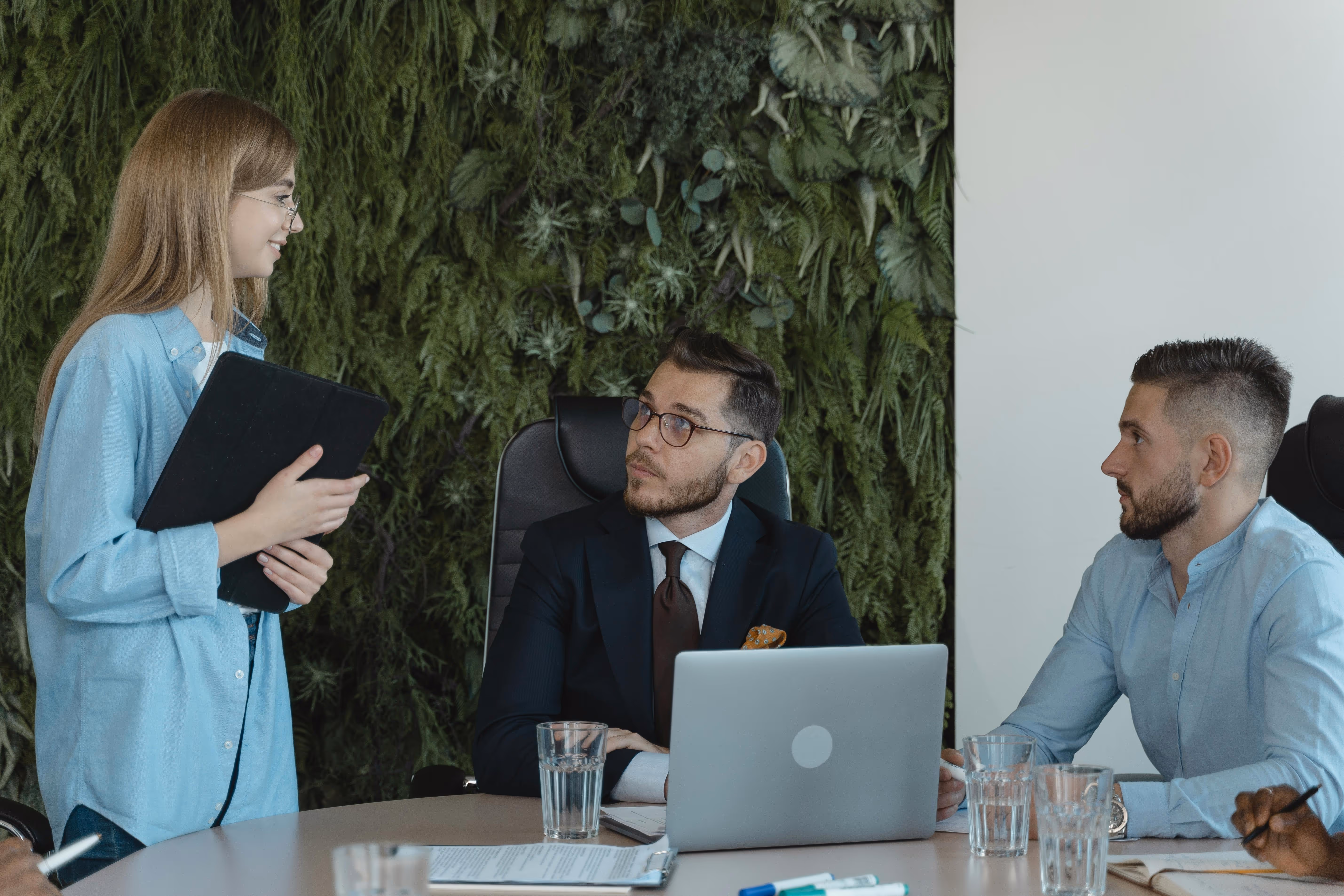 Three colleagues in a meeting room with a green plant wall, one woman standing with a tablet and two men seated at a table with a laptop and documents.